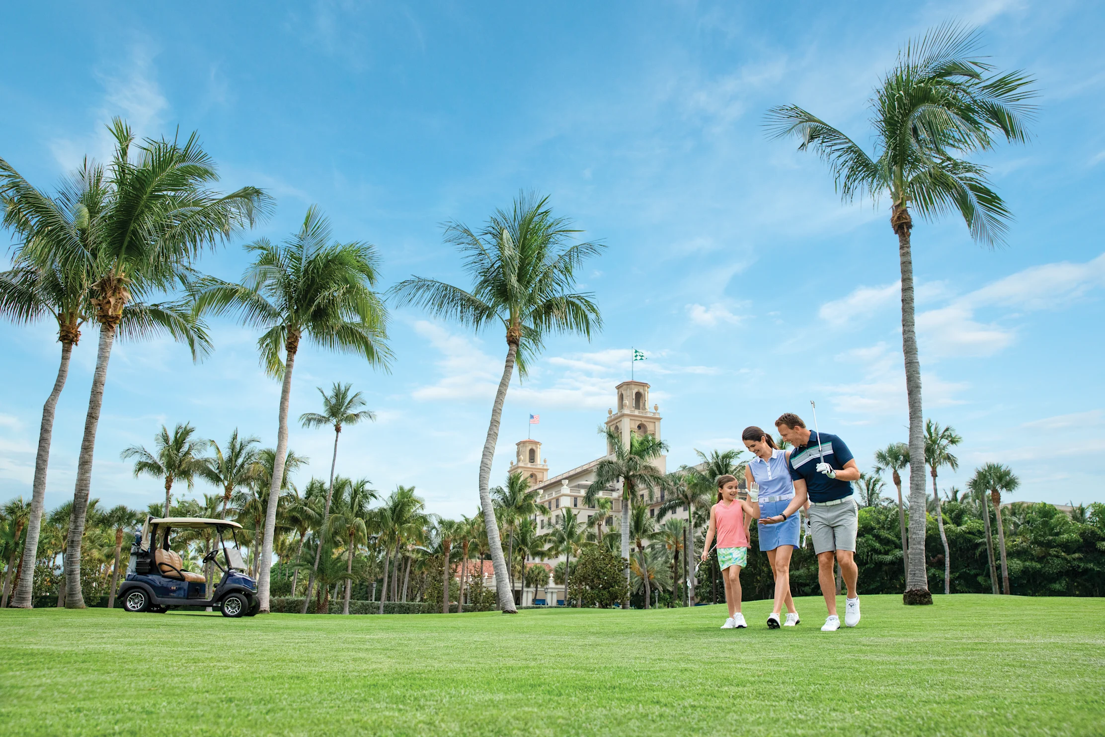 Family of three walking on golf course with palm trees and historic building under blue sky.