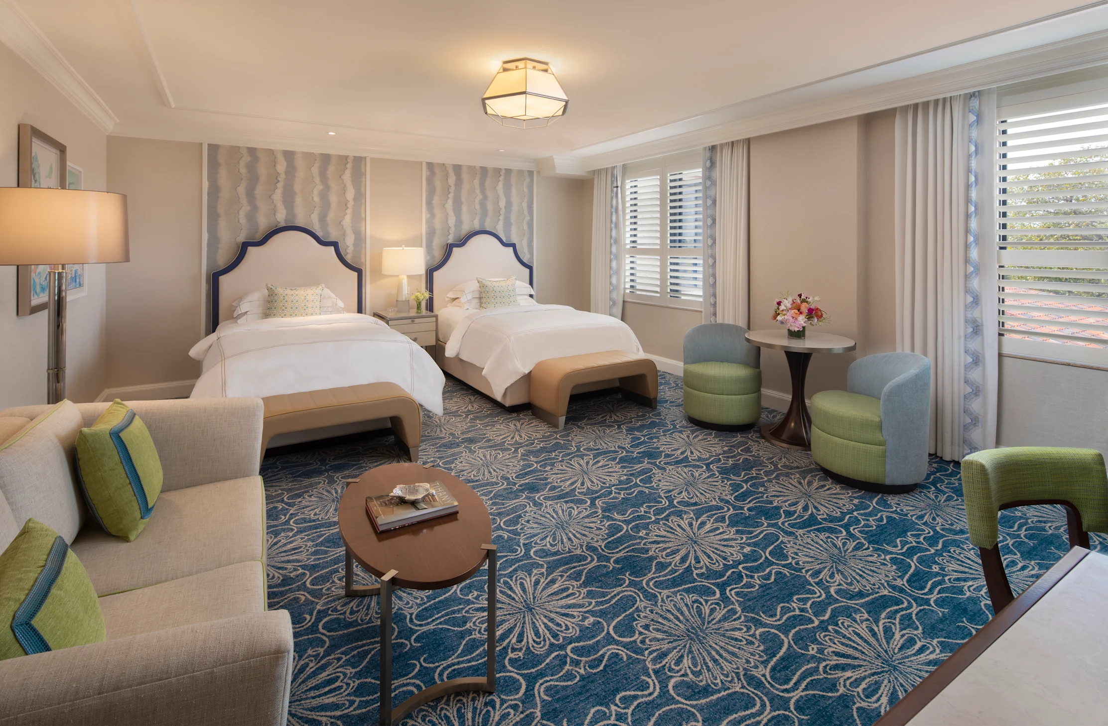 Elegant hotel room with two beds featuring navy headboards, blue patterned carpet, and green seating.