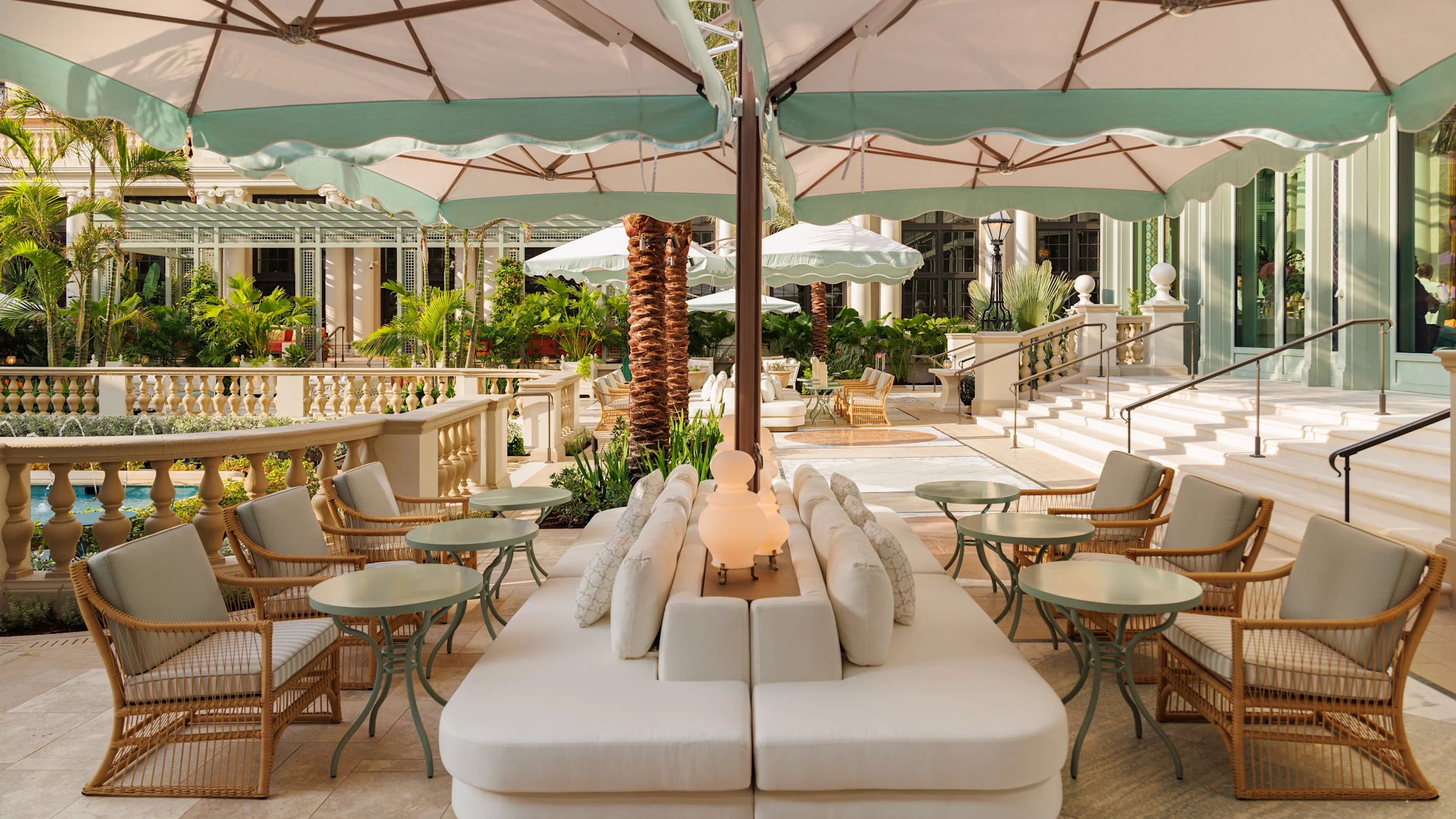 Elegant outdoor patio with white umbrellas, cream seating, wicker chairs, and tropical plants.