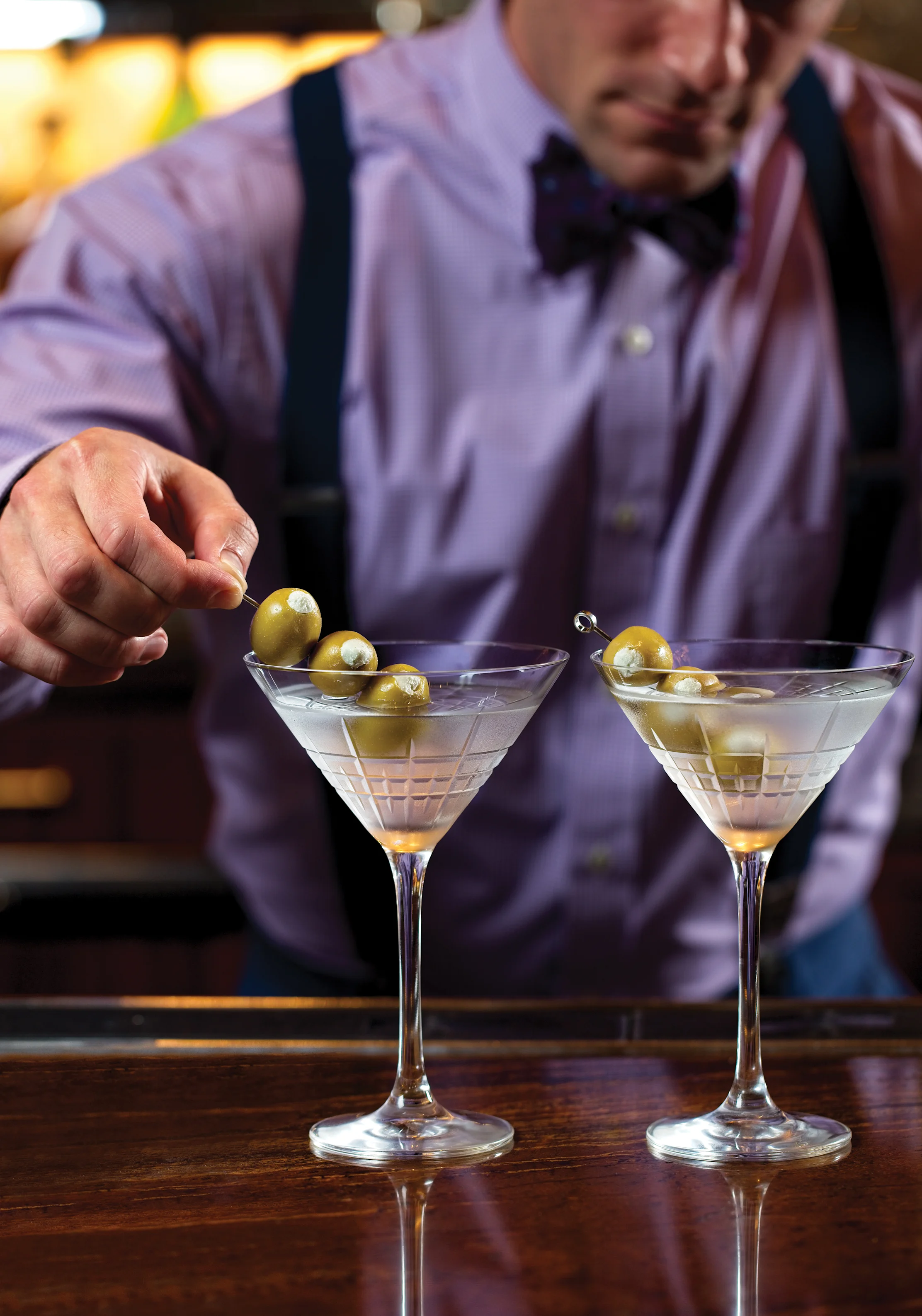 Bartender in purple shirt and bow tie garnishing two martinis with green olives on wooden bar.