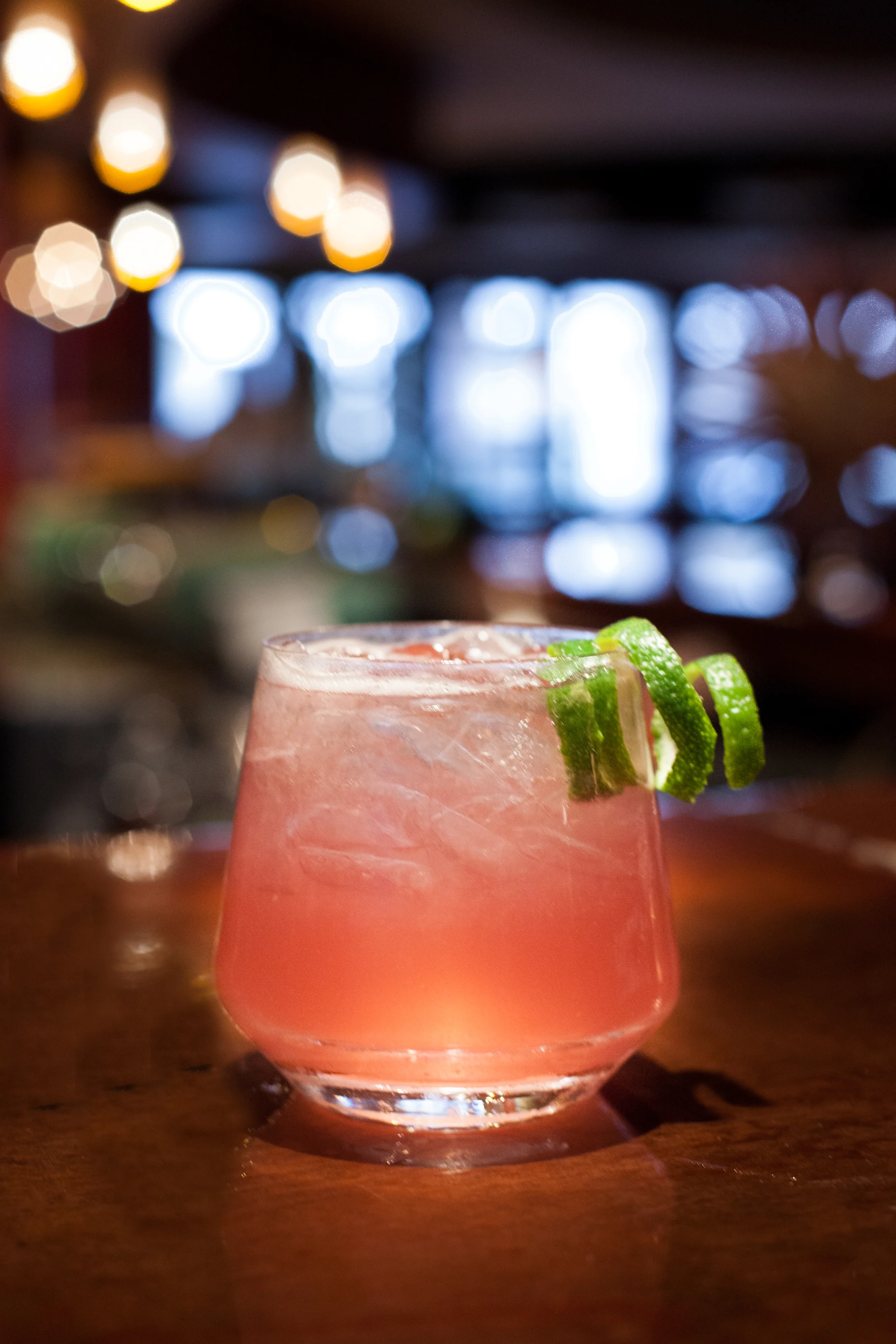 Pink cocktail with ice and lime garnish on a wooden bar with blurred lights in the background.