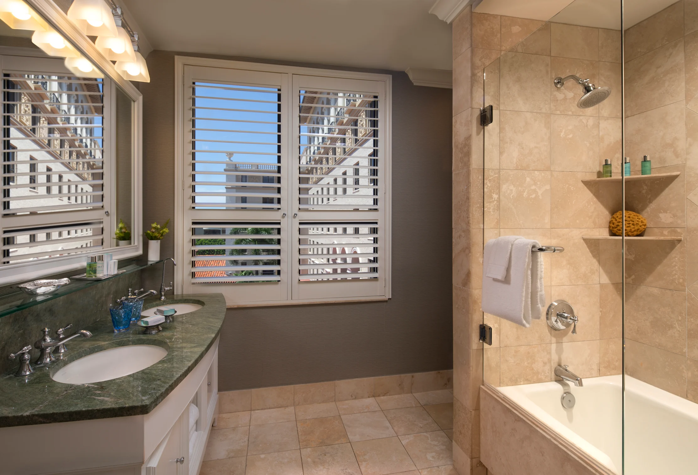 Modern bathroom with beige tile, glass shower, white shuttered windows, and green granite vanity.