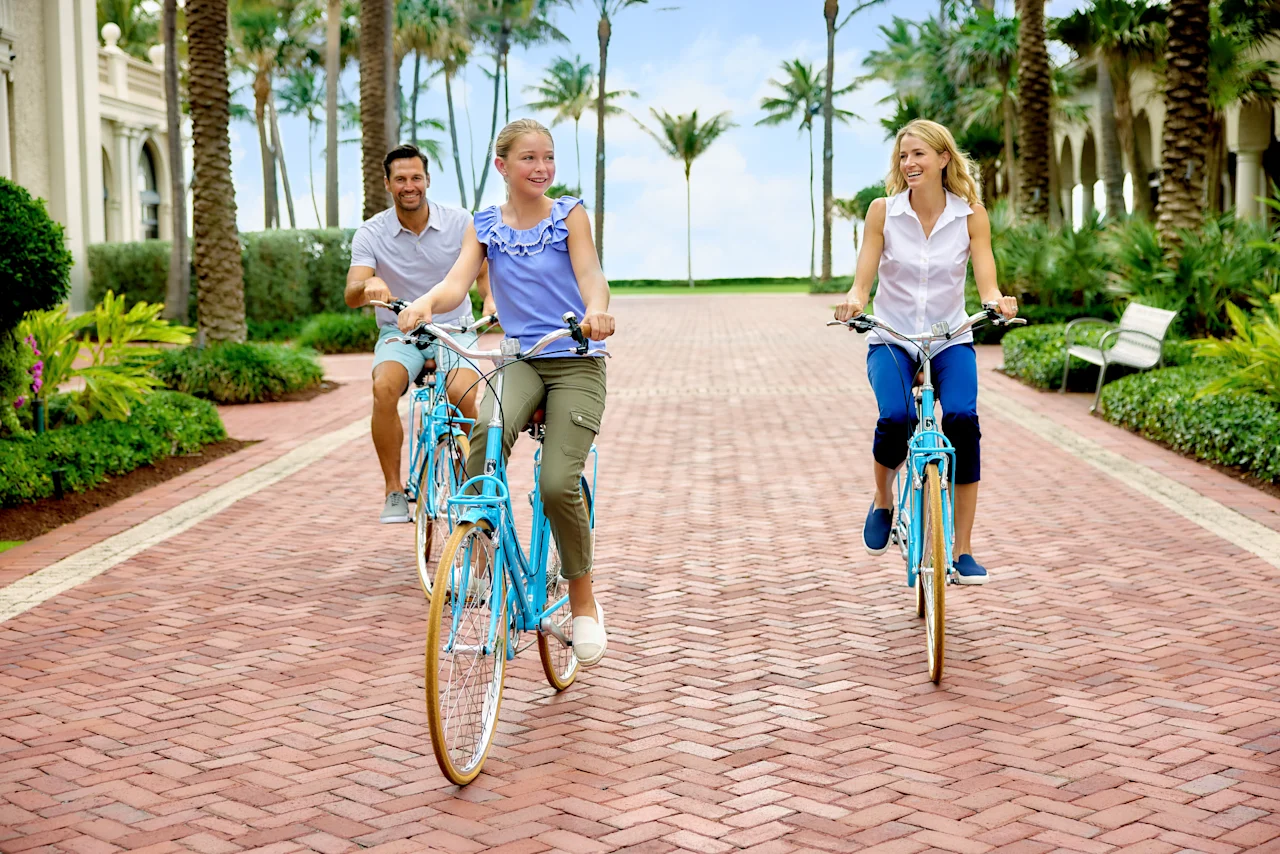 Family of three riding blue bicycles on brick path lined with palm trees at tropical resort.