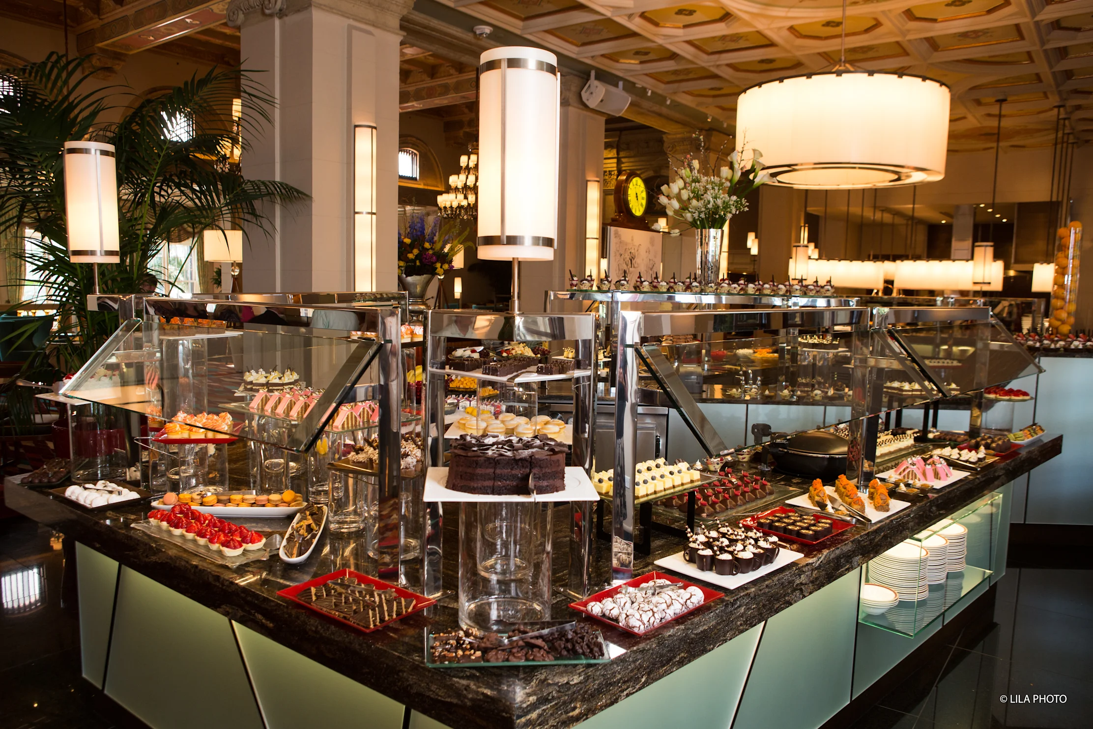 Elegant hotel buffet with tiered dessert displays, glass cases, modern lighting, and ornate ceiling.