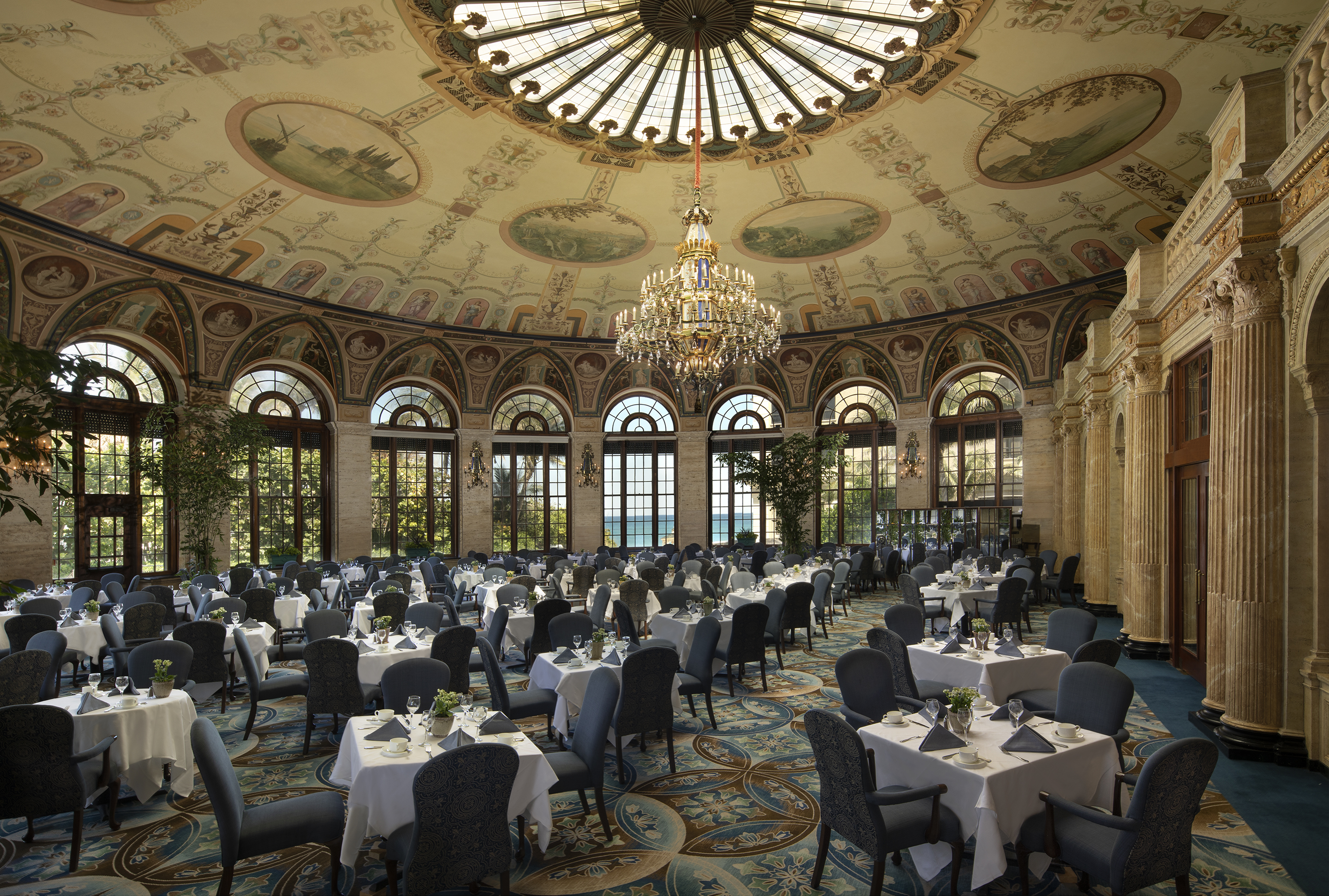 Elegant dining room with ornate painted ceiling, crystal chandelier, arched windows, and tables set with white linens.
