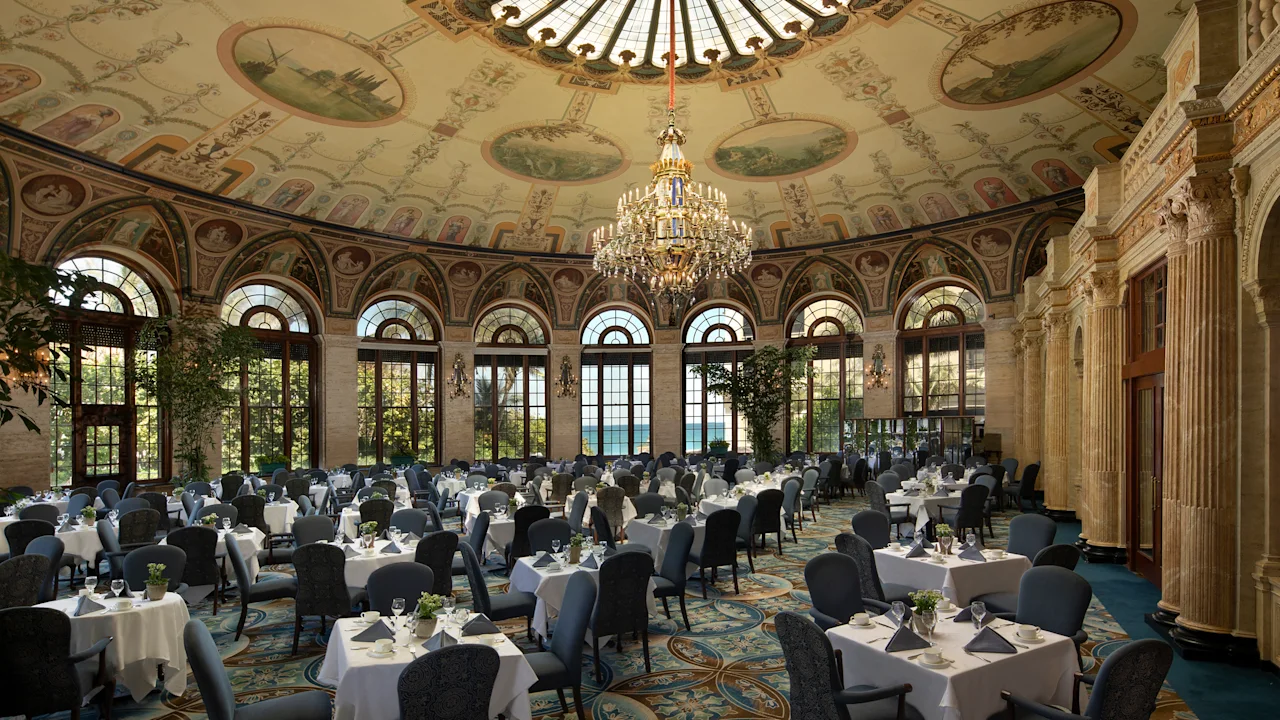 Elegant dining room with ornate painted ceiling, crystal chandelier, arched windows, and tables set with white linens.