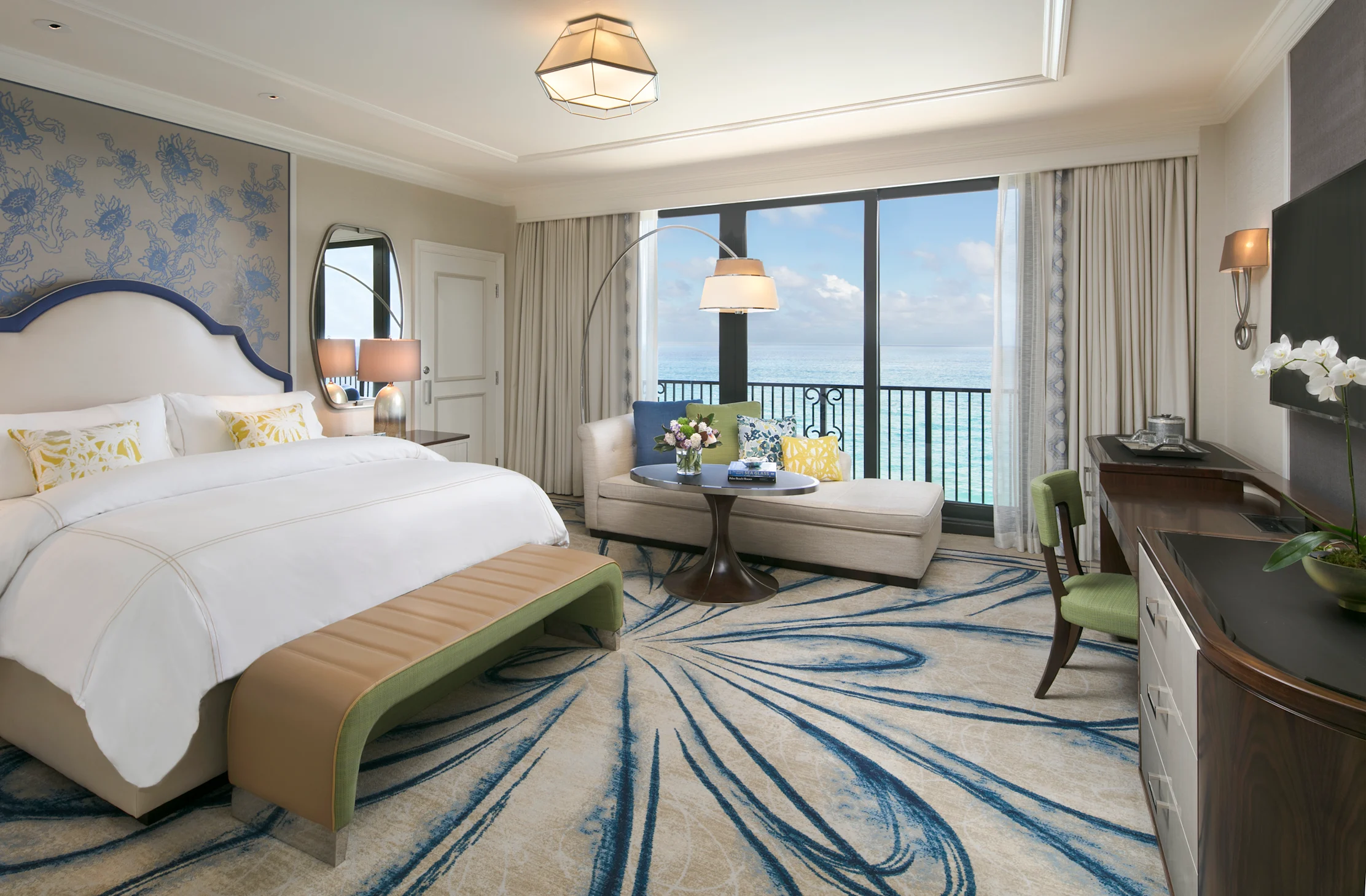 Elegant hotel room with ocean view, white bed, blue accents, seating area, and balcony overlooking water.