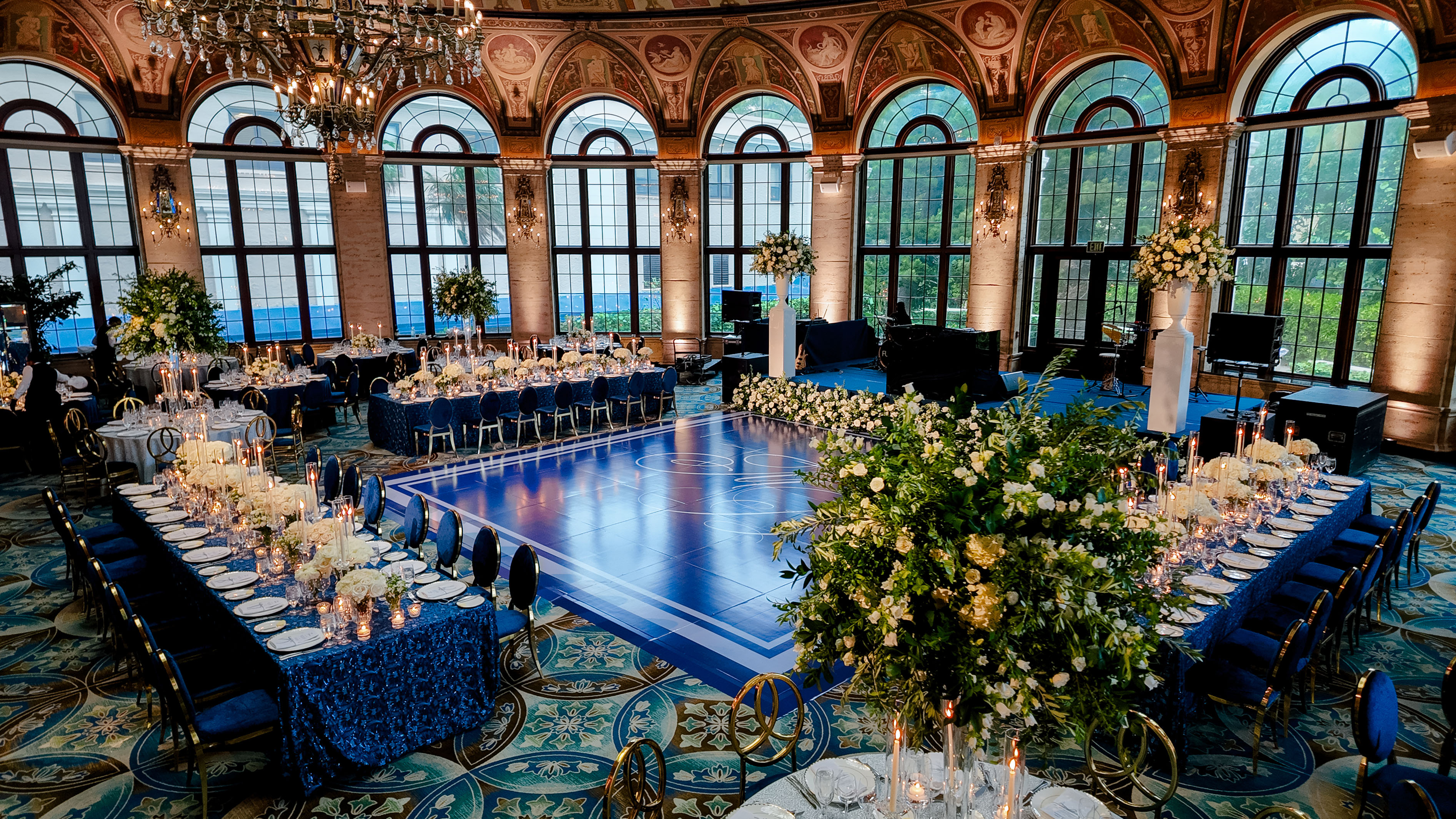 Elegant ballroom with arched windows, chandeliers, blue dance floor, and formal dining tables.