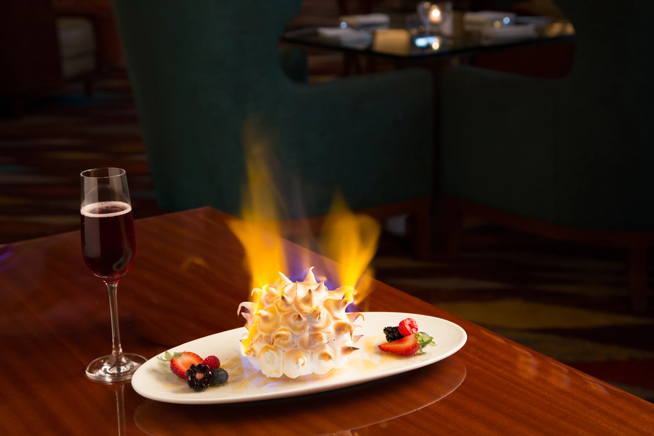 Flaming Baked Alaska dessert with berries on white plate beside glass of red wine on wooden table