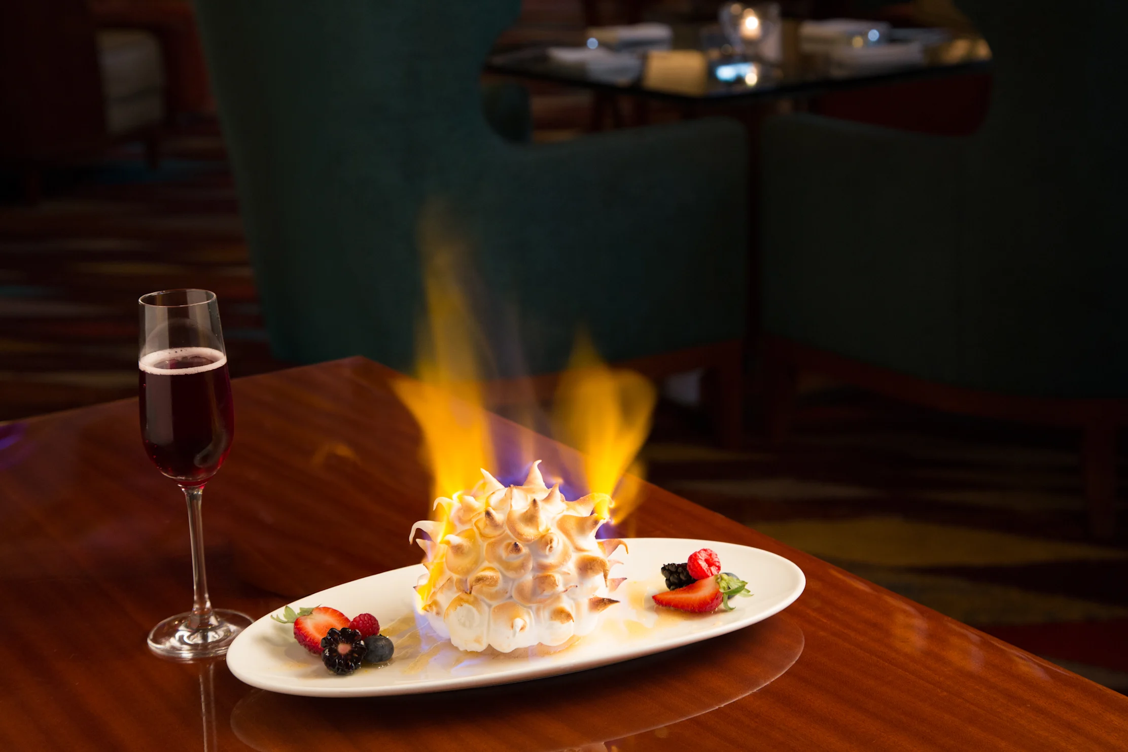 Flaming Baked Alaska dessert with berries on white plate beside glass of red wine on wooden table