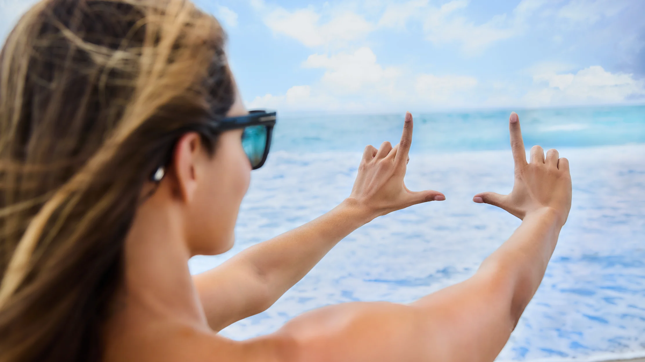 Woman with sunglasses framing ocean view with hands against blue sky and sea background.