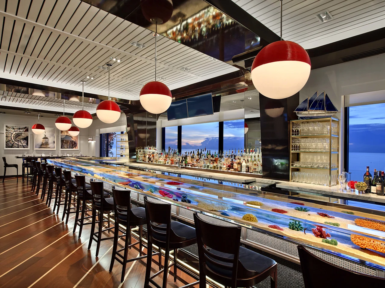 Modern bar with red pendant lights, illuminated counter with colorful display, and ocean view windows.