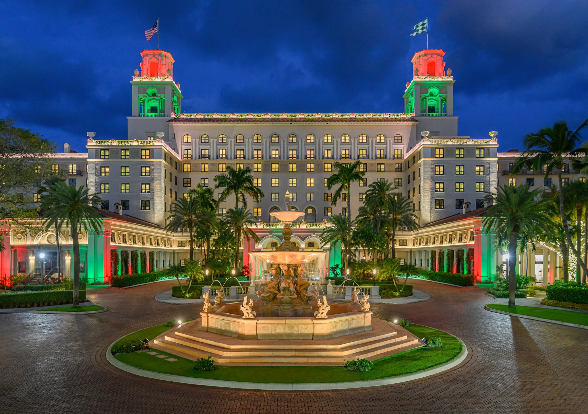 Grand hotel with red and green illuminated towers, fountain courtyard, and palm trees at dusk.