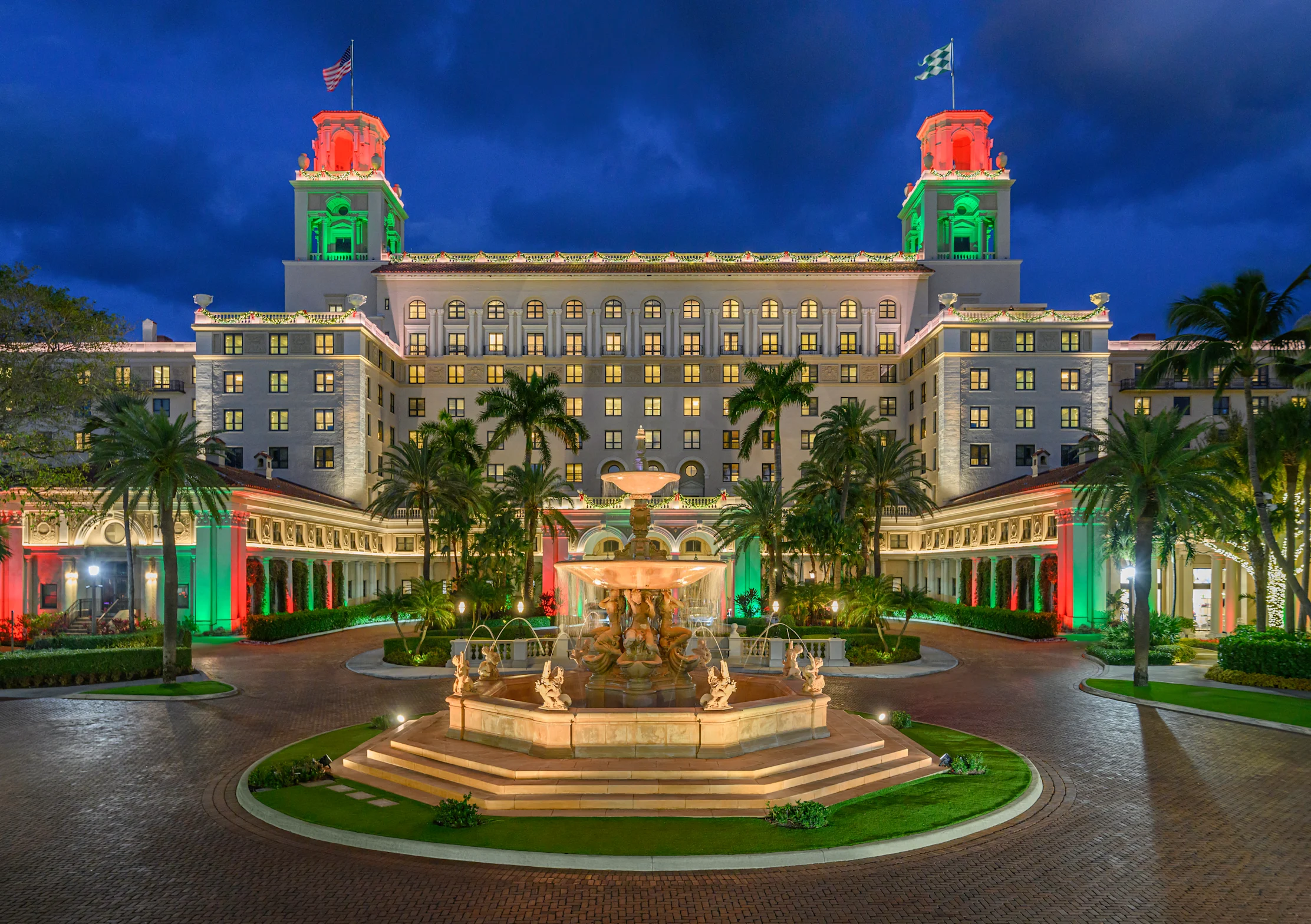 Grand hotel with red and green illuminated towers, fountain courtyard, and palm trees at dusk.