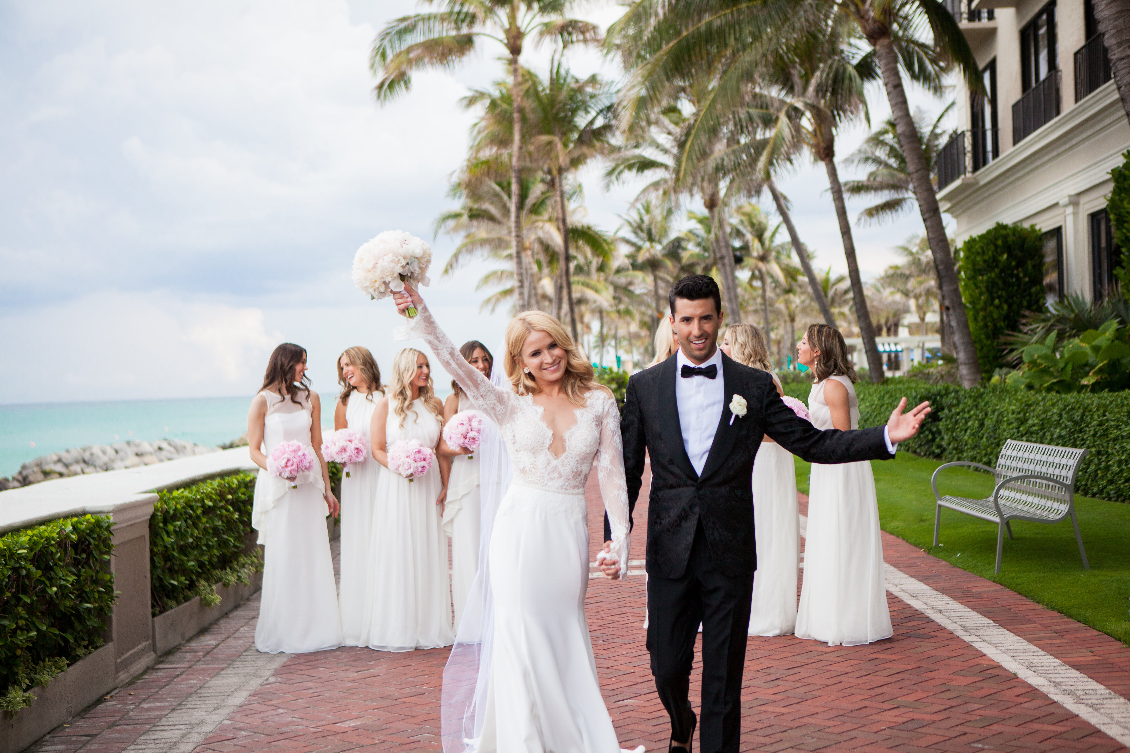 Bride and groom celebrating with bridesmaids at tropical beachfront wedding venue with palm trees.