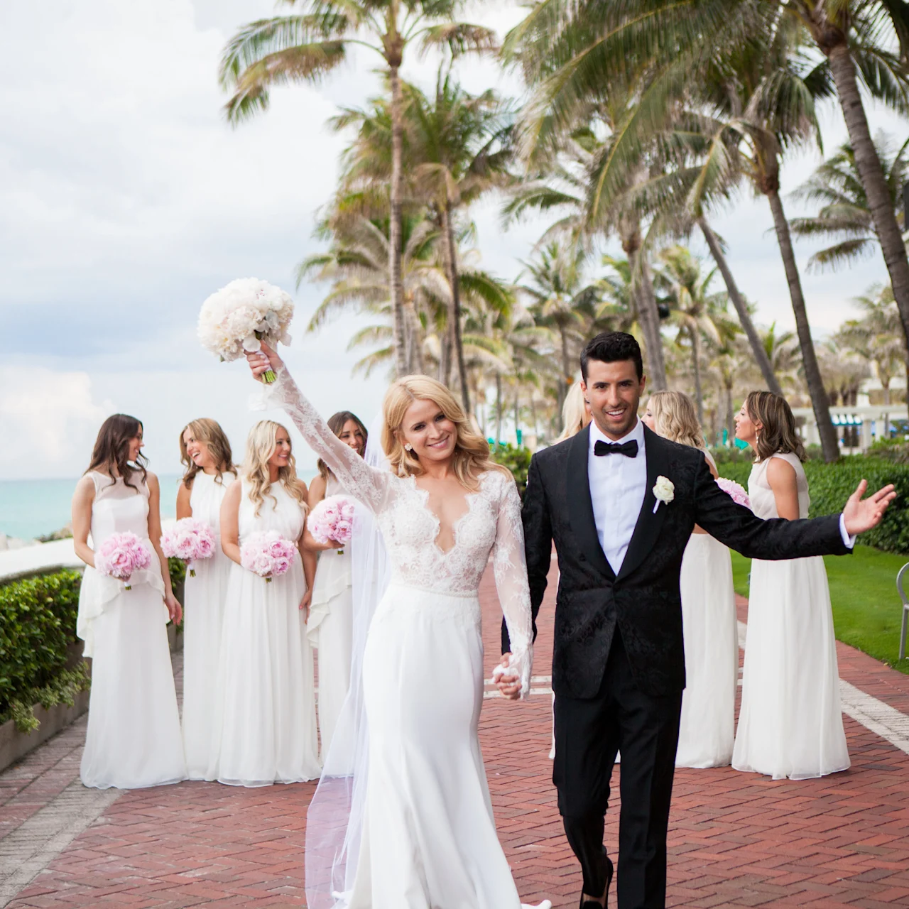 Bride and groom celebrating with bridesmaids at tropical beachfront wedding venue with palm trees.