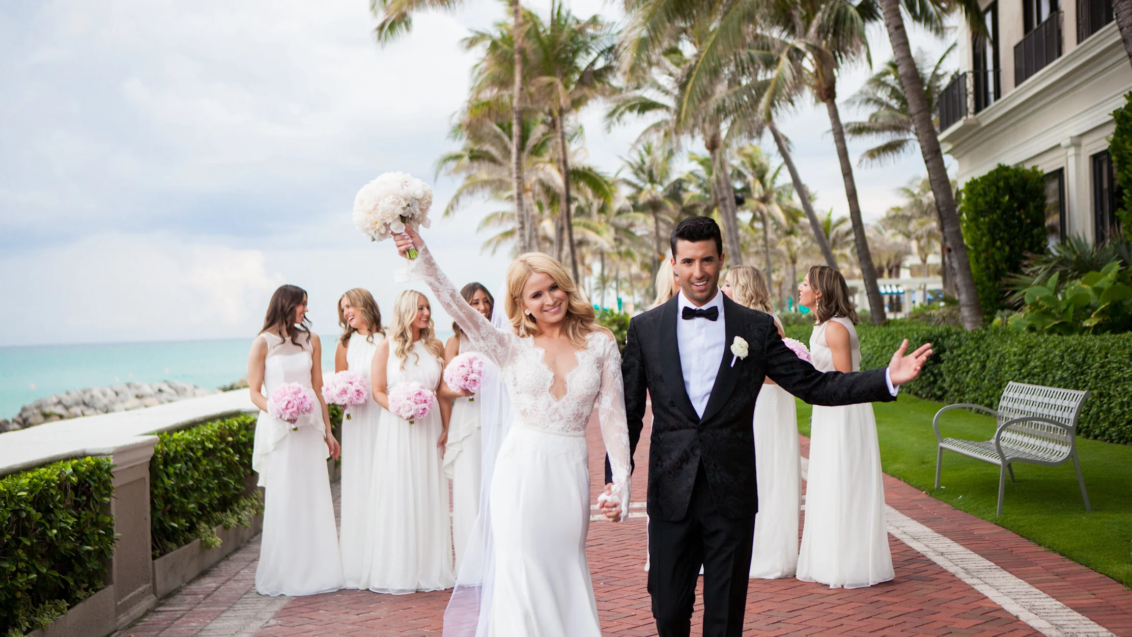Bride and groom celebrate with bridesmaids at tropical beachfront wedding venue with palm trees.