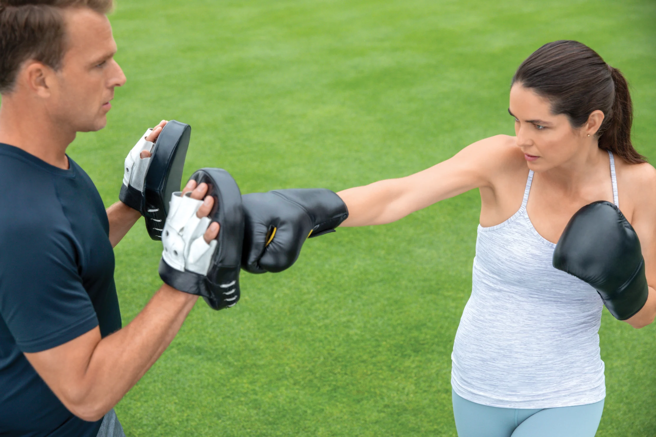 Woman in boxing gloves training with male coach holding pads outdoors on green grass field.