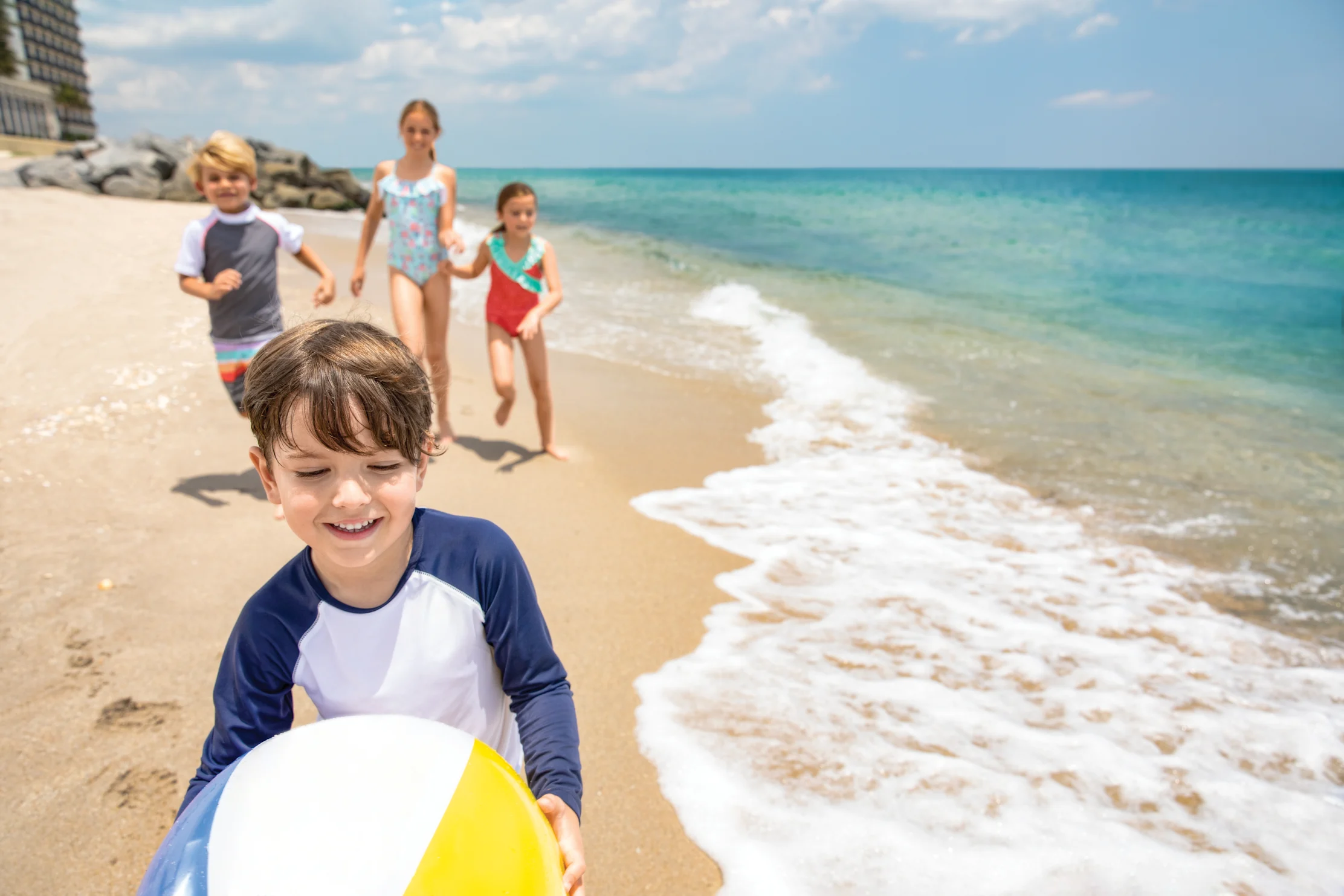 Four children running and playing on a sunny beach with turquoise water and white waves.