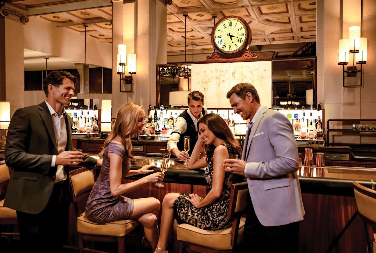 Elegantly dressed guests enjoying cocktails at an upscale hotel bar with ornate ceiling and vintage clock.