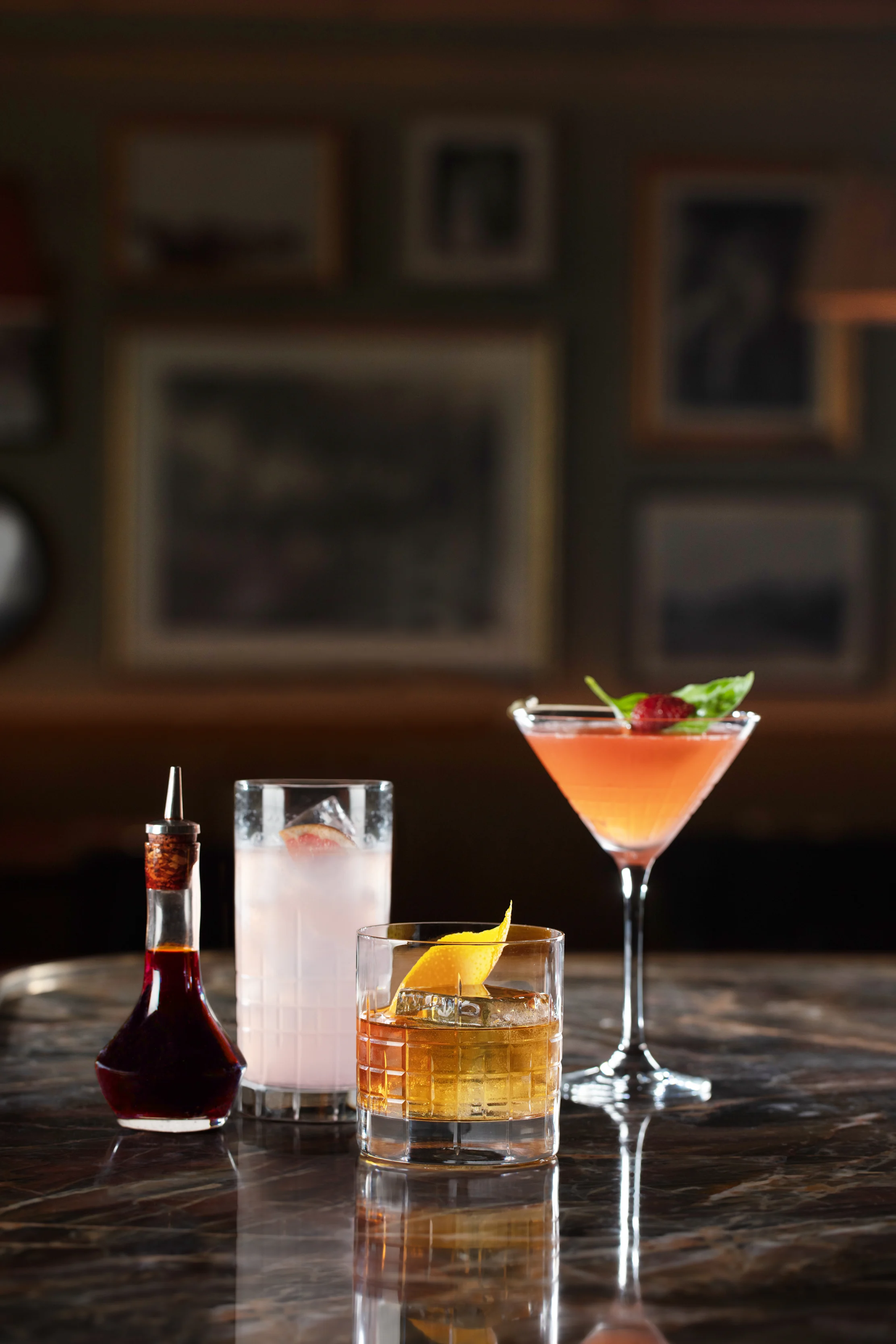Four cocktails displayed on a dark wooden bar with framed artwork in the blurred background.