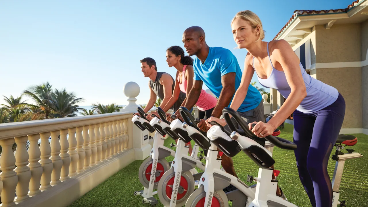 Four people exercising on stationary bikes outdoors on a terrace with palm trees and blue sky.