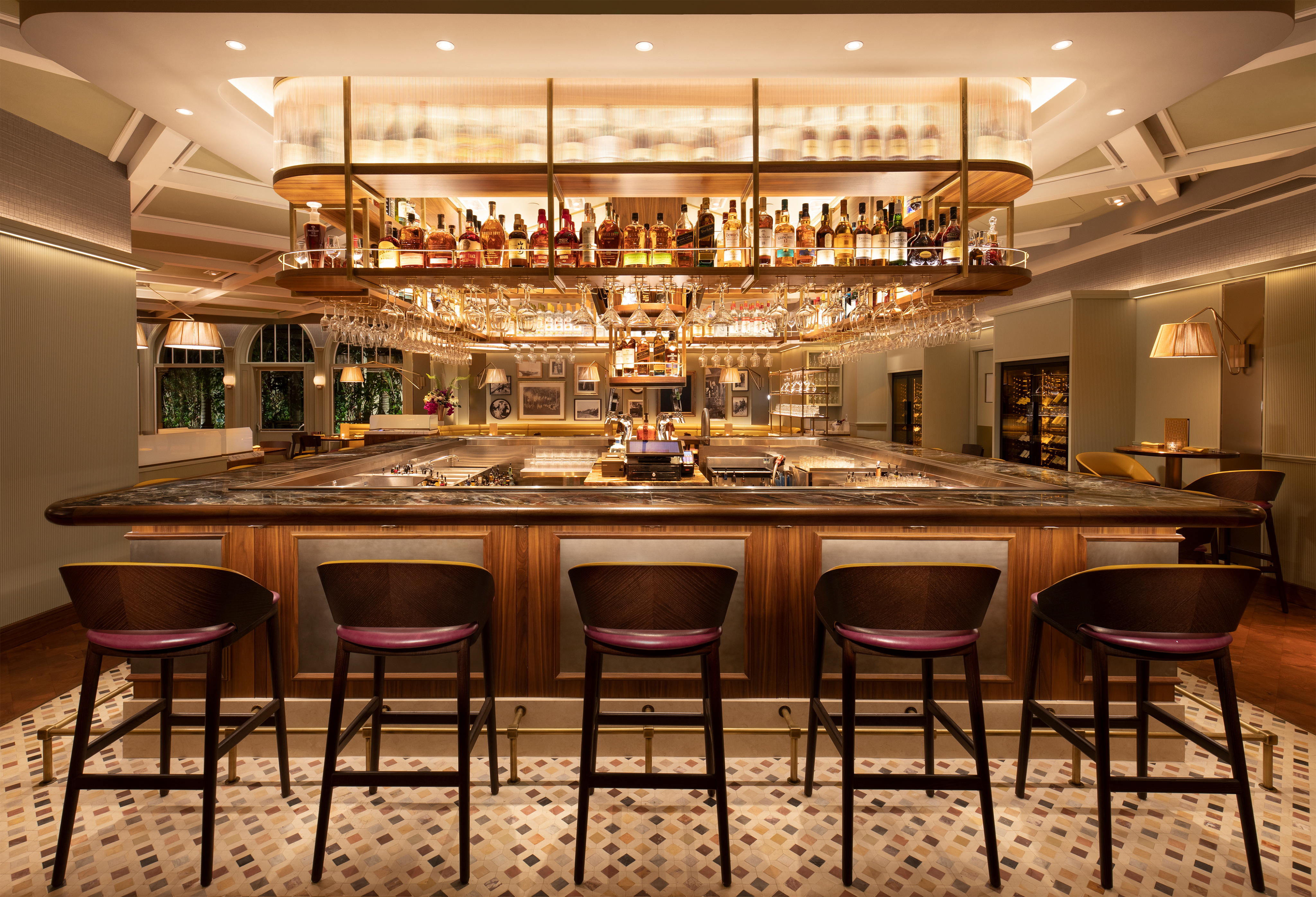 Upscale hotel bar with wooden counter, leather bar stools, and illuminated liquor display overhead.