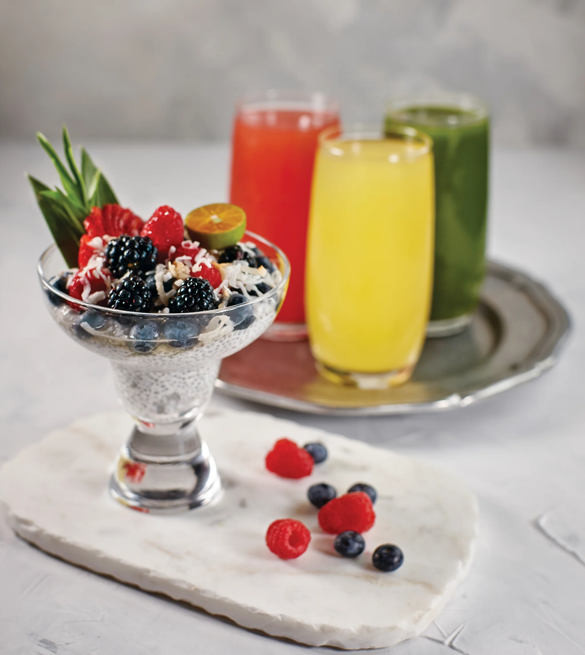 Chia pudding with berries in glass bowl alongside three colorful smoothies in red, yellow, and green.