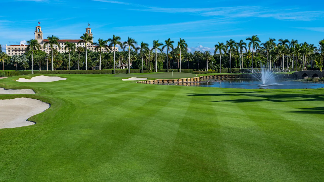 Lush green golf course with sand bunkers, water fountain, palm trees, and resort building under blue sky.
