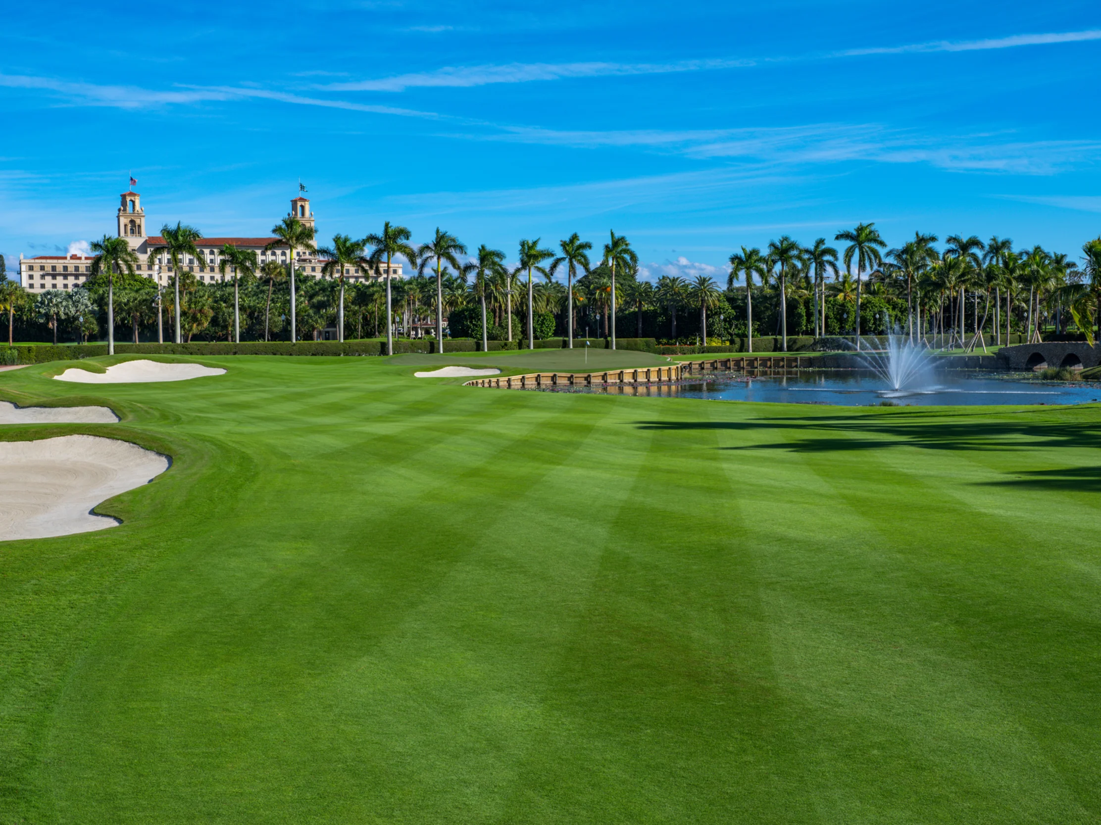 Lush green golf course with sand bunkers, water fountain, palm trees, and resort building under blue sky.