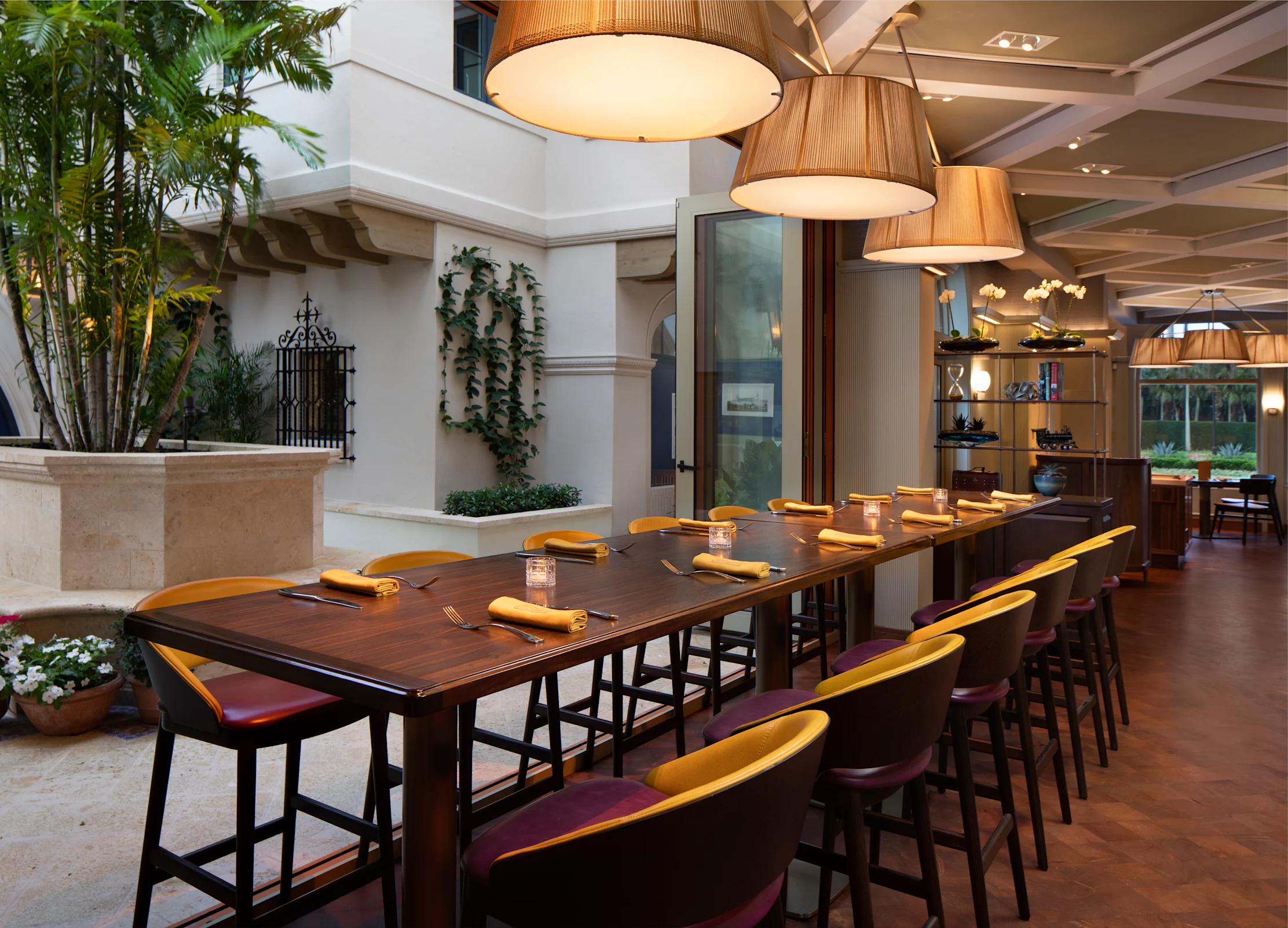 Elegant restaurant dining area with long dark wood table, yellow napkins, hanging pendant lights, and lush greenery.
