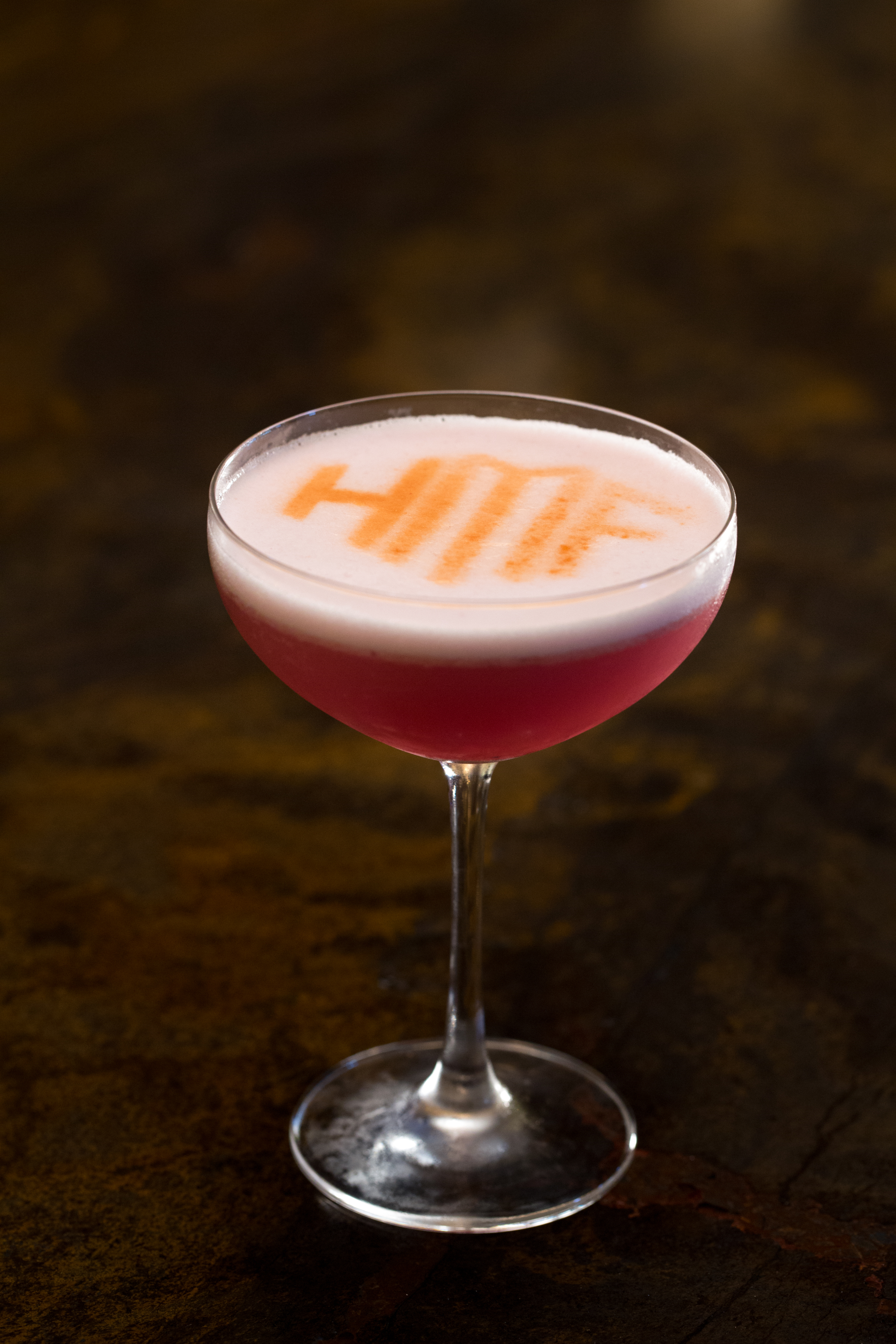 Pink cocktail in coupe glass with white foam top and orange text design on dark background