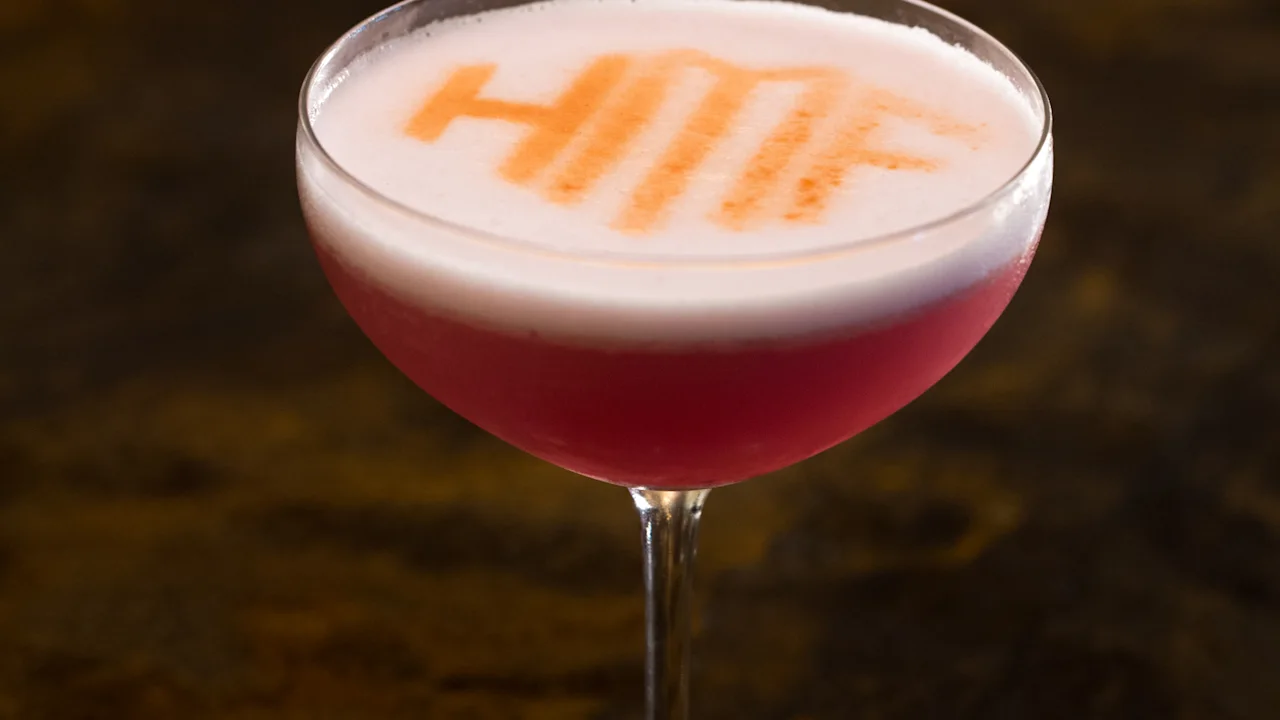 Pink cocktail in coupe glass with white foam top and orange text design on dark background