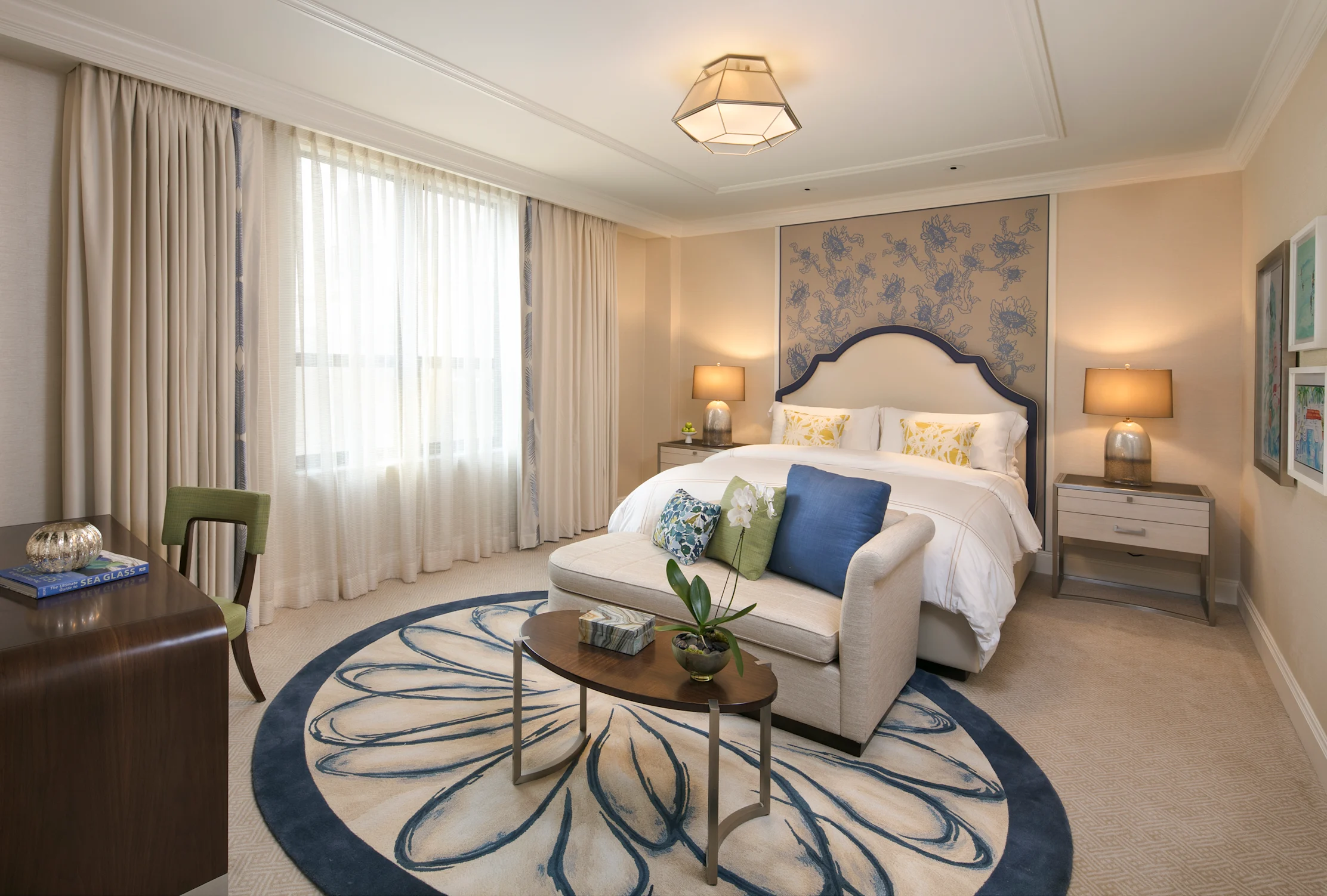 Elegant hotel bedroom with white bed, blue headboard, beige curtains, seating area, and geometric ceiling light.