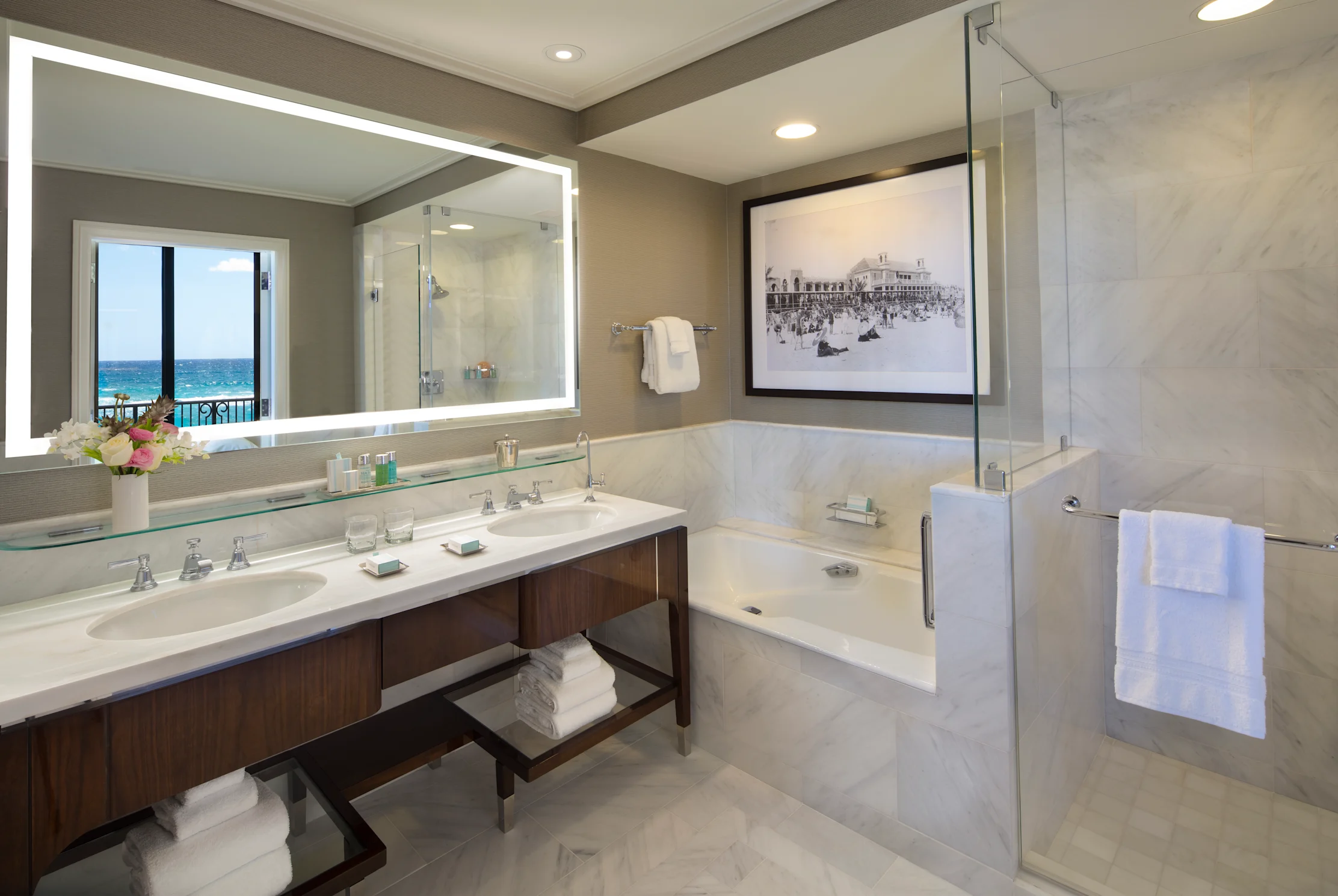 Modern luxury bathroom with double vanity, soaking tub, glass shower, and ocean view through mirror.