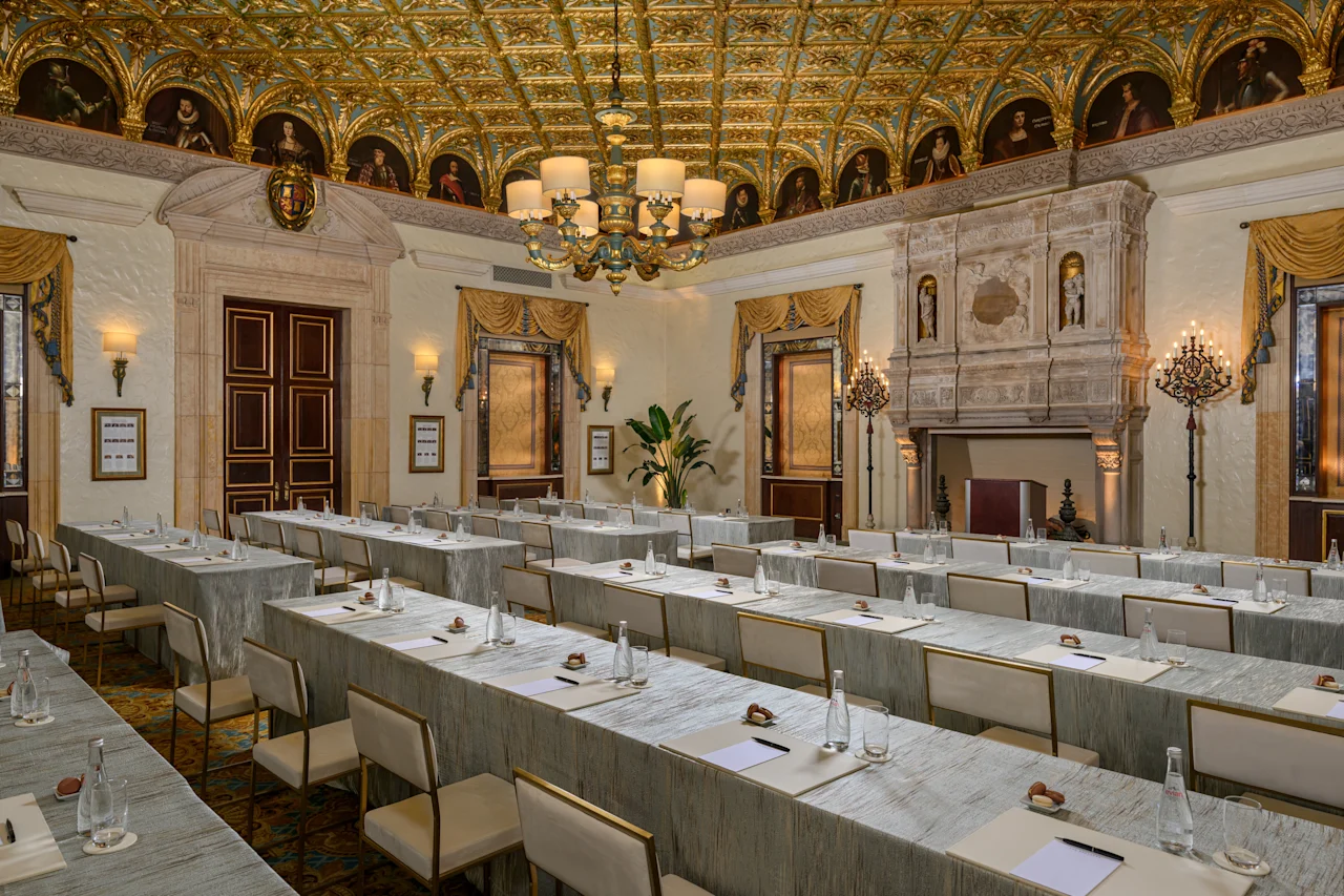 Ornate conference room with gold-decorated ceiling, chandelier, marble walls, and rows of tables.