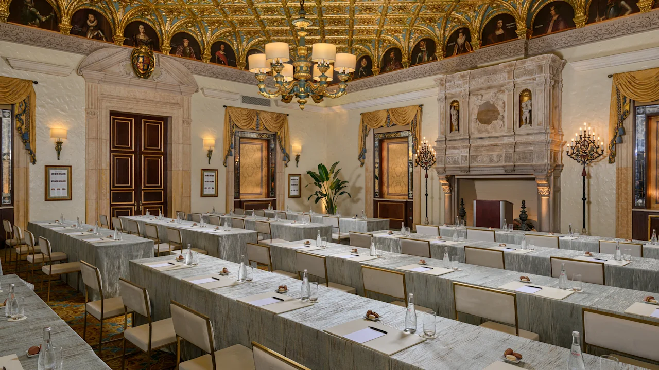 Ornate conference room with gold-decorated ceiling, chandelier, marble walls, and rows of tables.