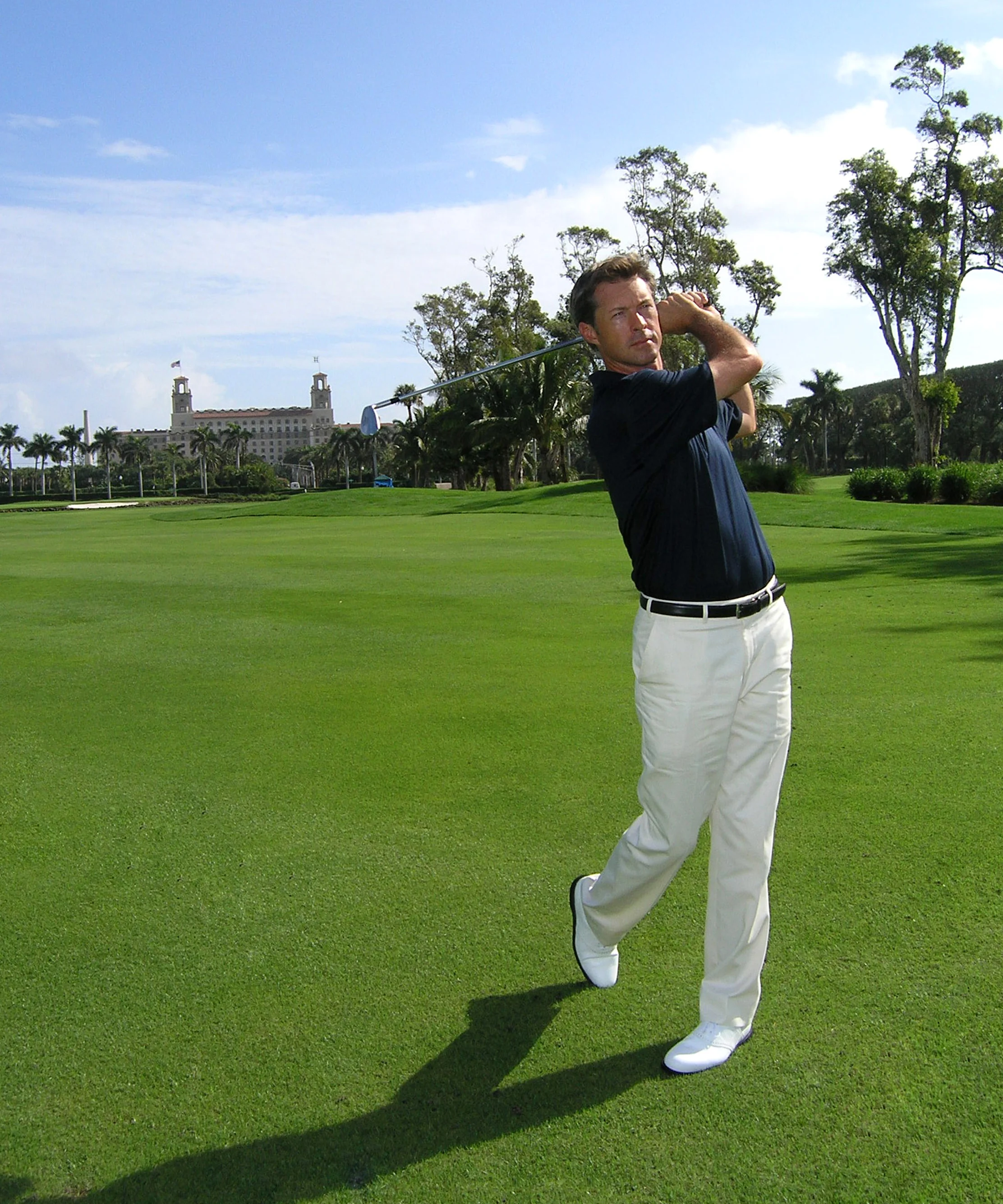 Man in navy shirt and white pants swinging golf club on green course with resort building in background.