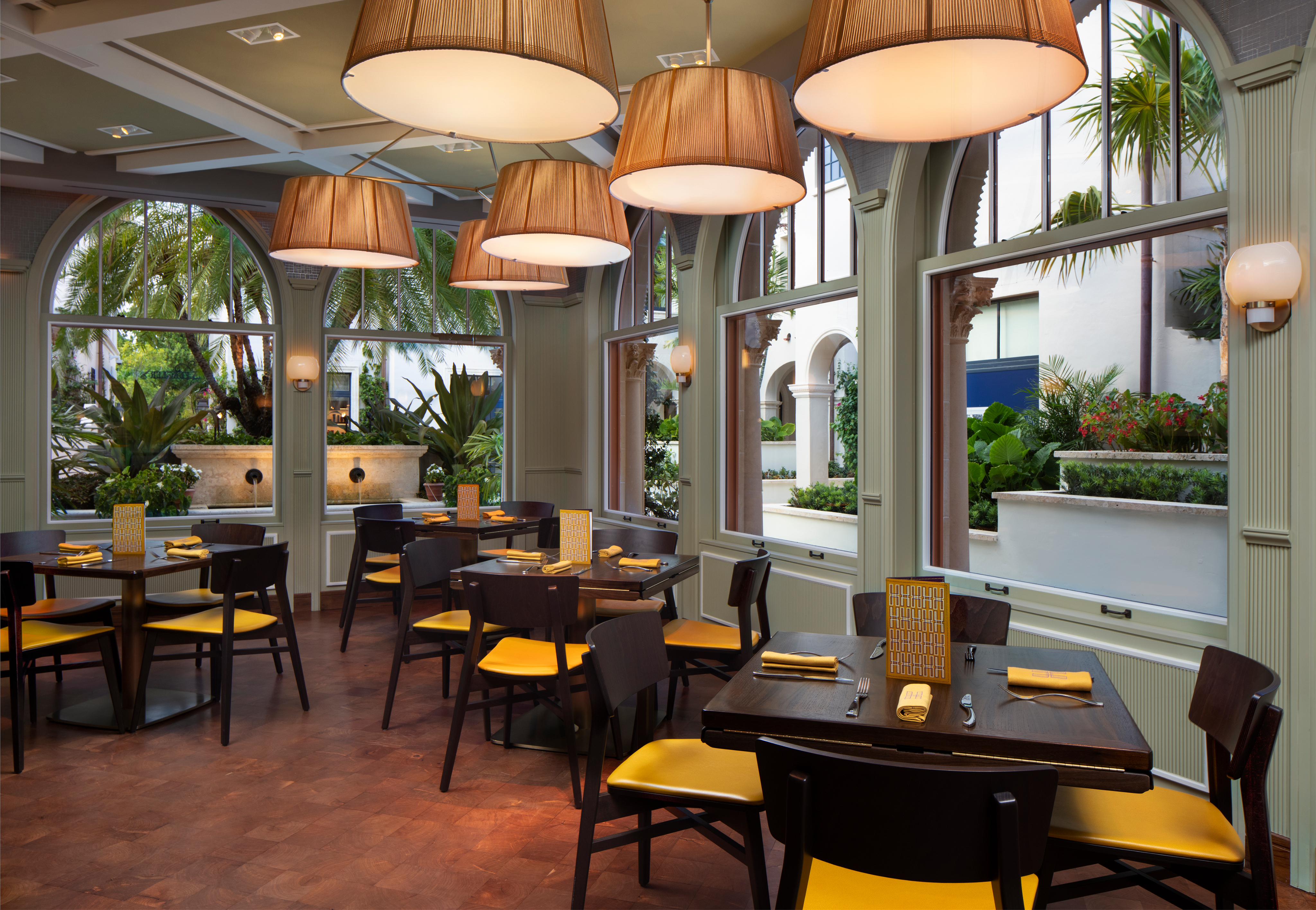 Elegant restaurant interior with dark wood tables, yellow chairs, tan pendant lights, and windows overlooking tropical courtyard.