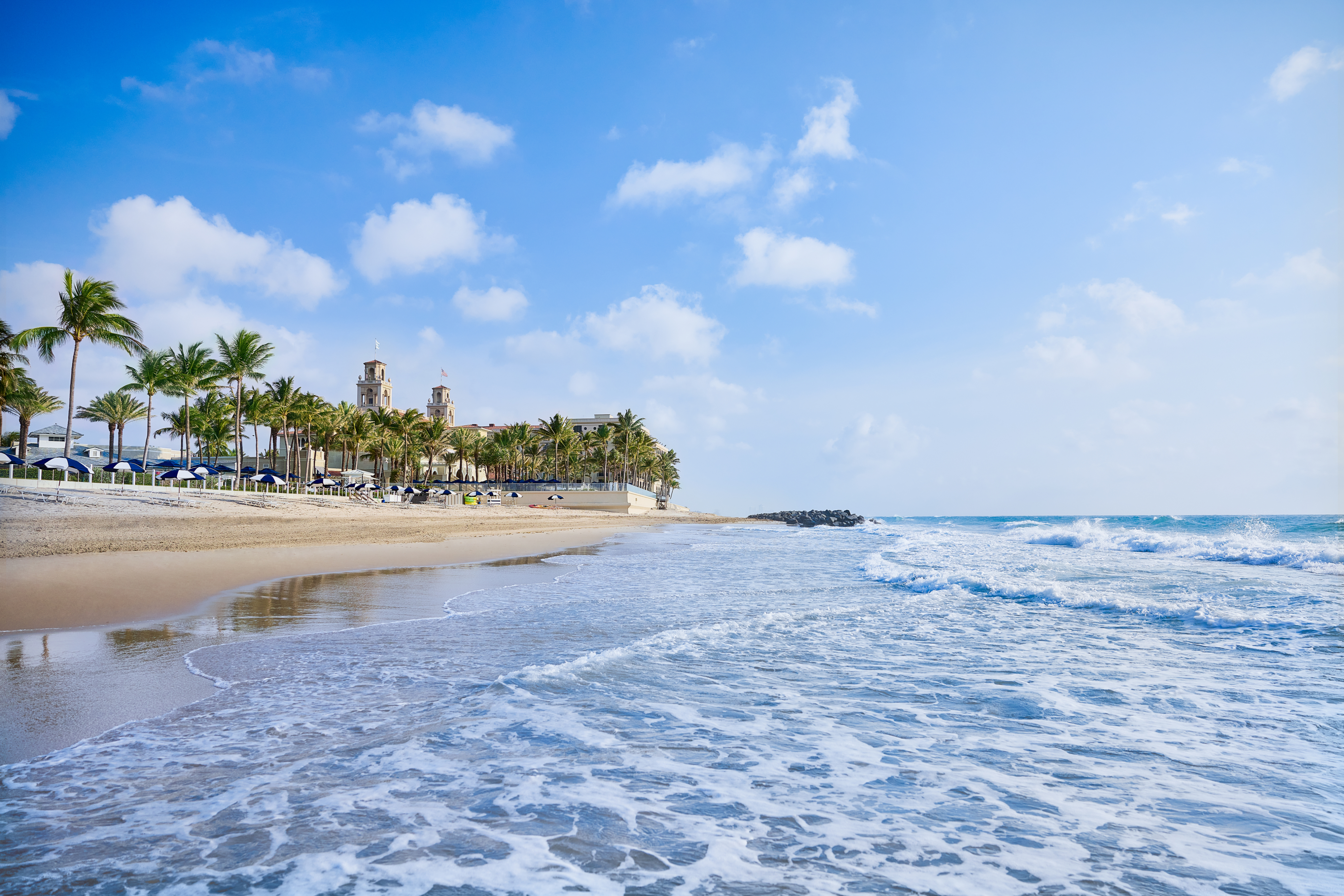Sandy beach with palm trees and historic building under blue sky with white clouds and ocean waves.