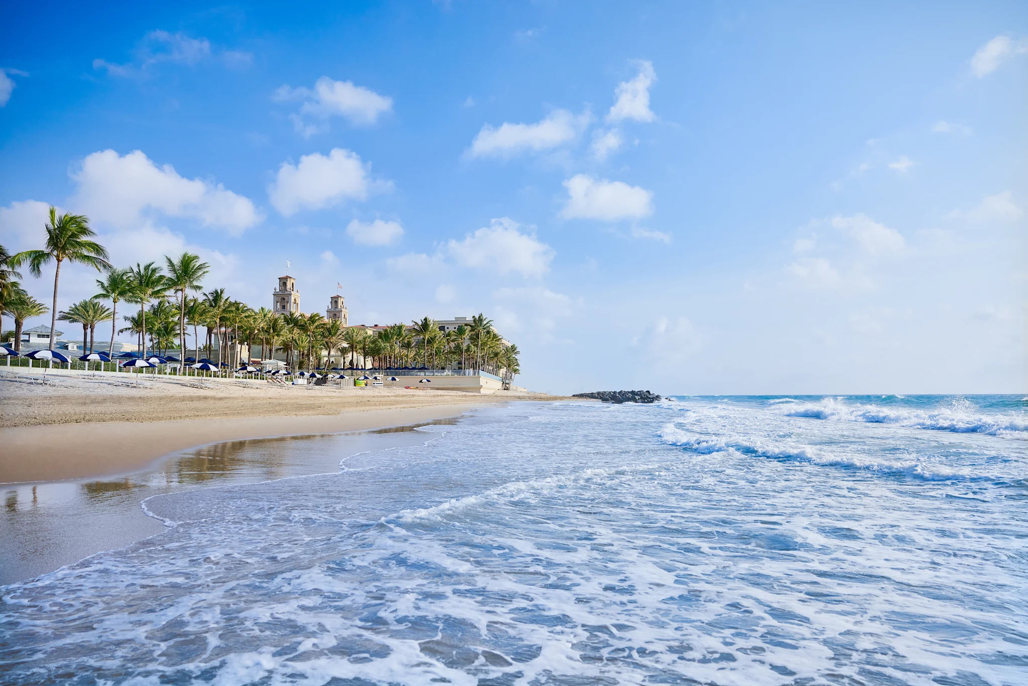 Sandy beach with palm trees and historic building under blue sky with white clouds and ocean waves.