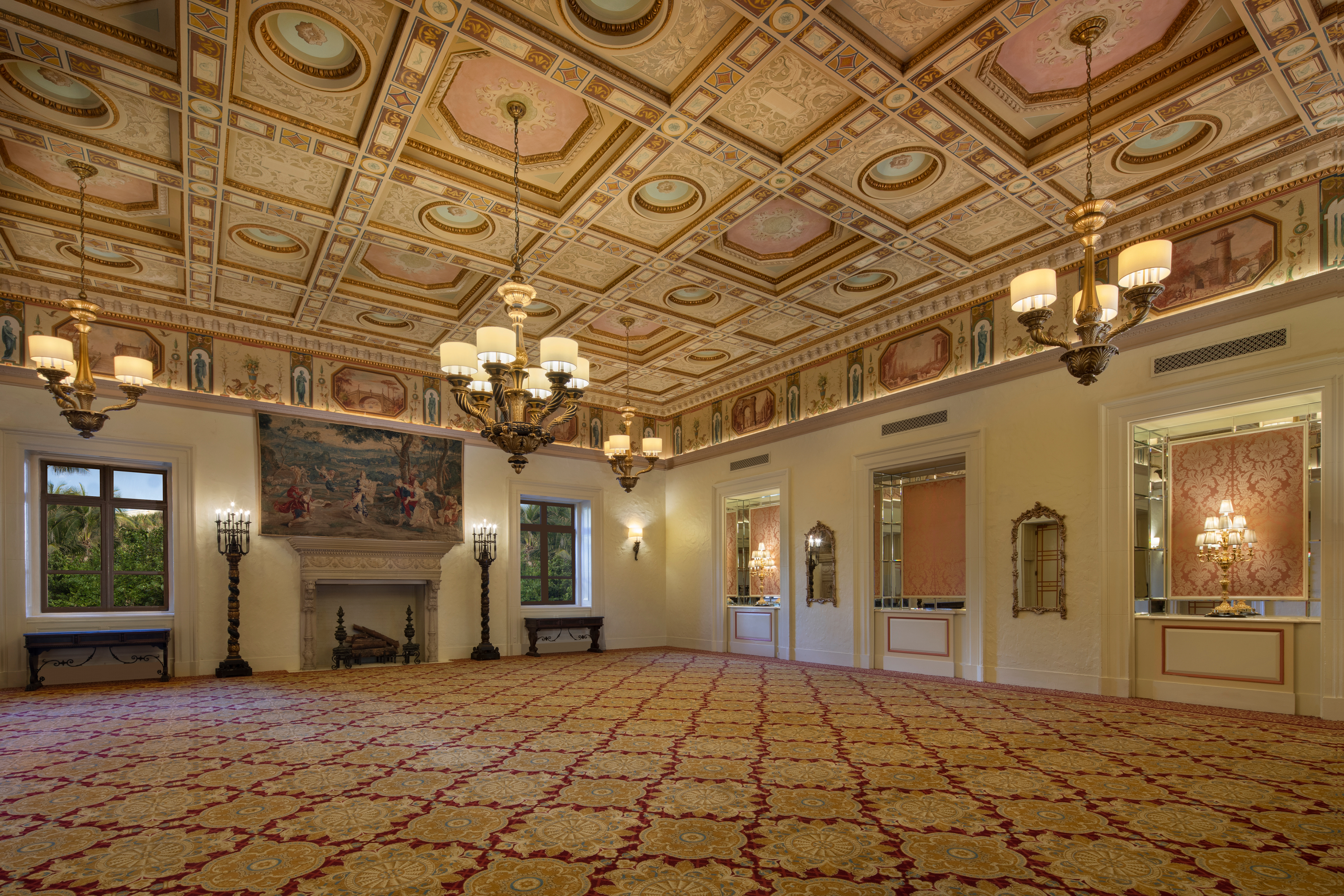 Ornate ballroom with decorative ceiling, chandeliers, fireplace, patterned carpet, and cream walls.