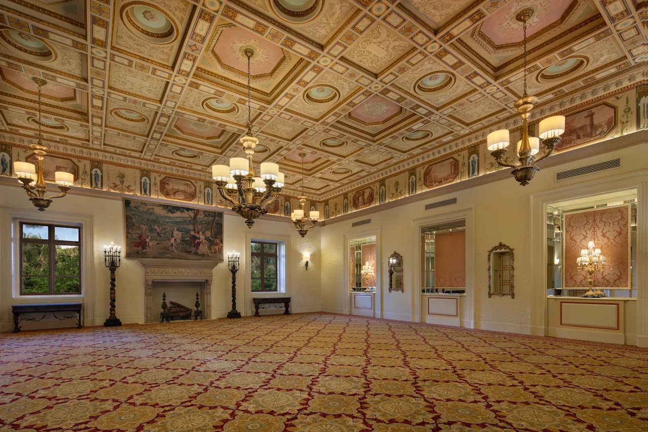 Ornate ballroom with decorative ceiling, chandeliers, fireplace, patterned carpet, and cream walls.
