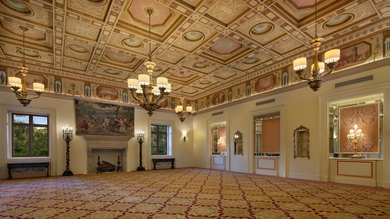 Ornate ballroom with decorative ceiling, chandeliers, fireplace, patterned carpet, and cream walls.