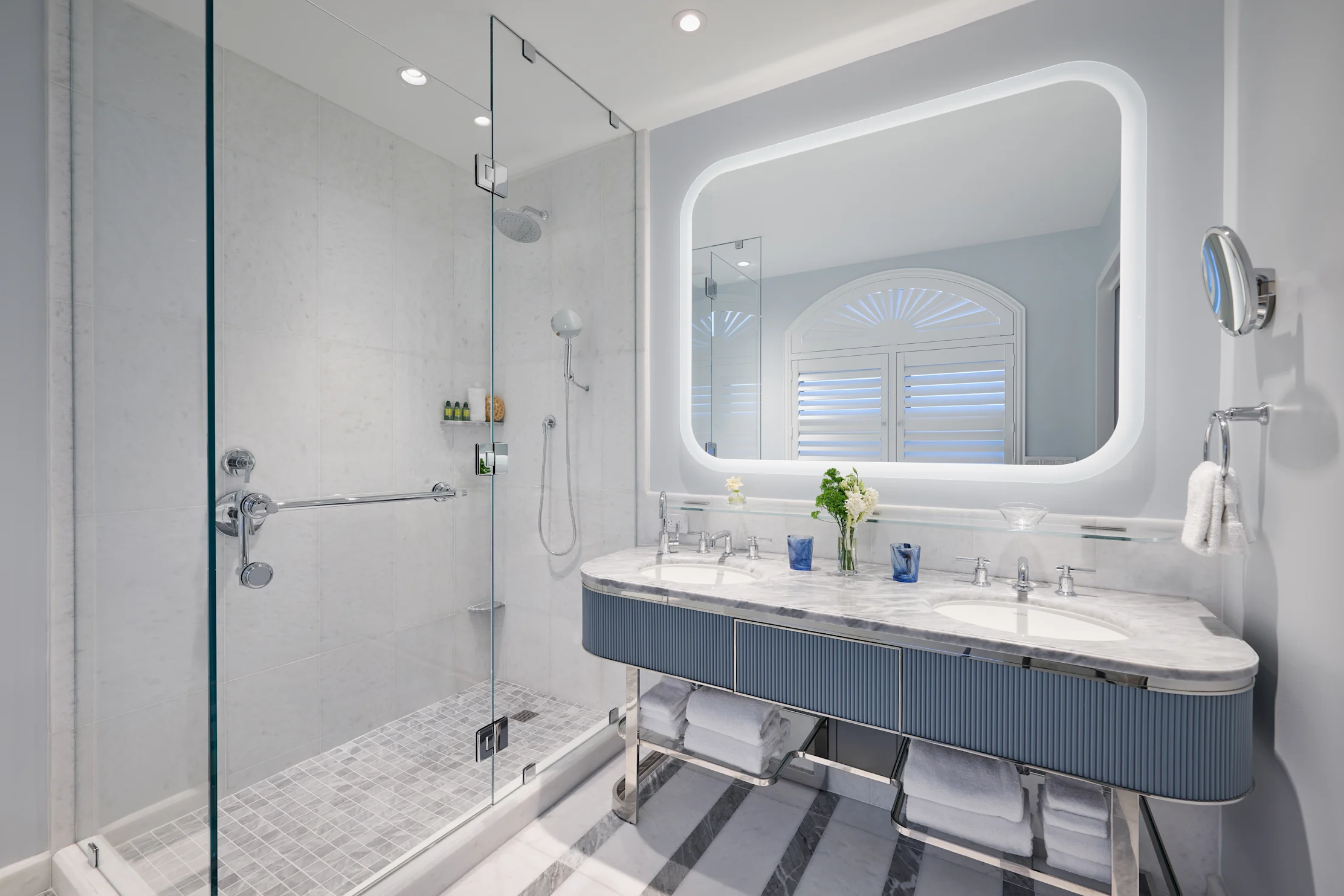 Modern bathroom with white marble vanity, blue accents, glass shower, and illuminated mirror.