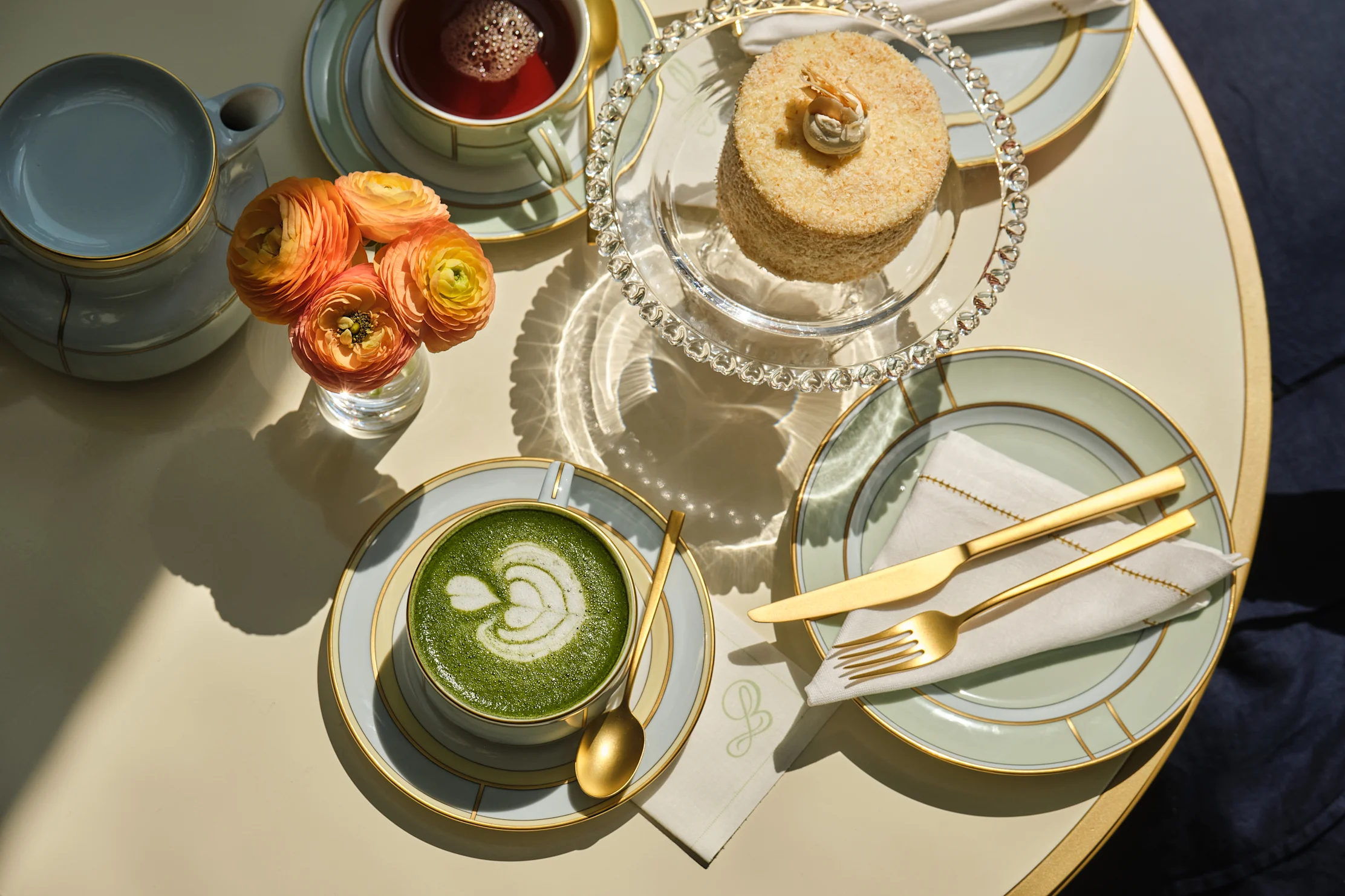 Elegant setup with matcha latte, tea, cake, orange flowers, and gold-trimmed china on white table