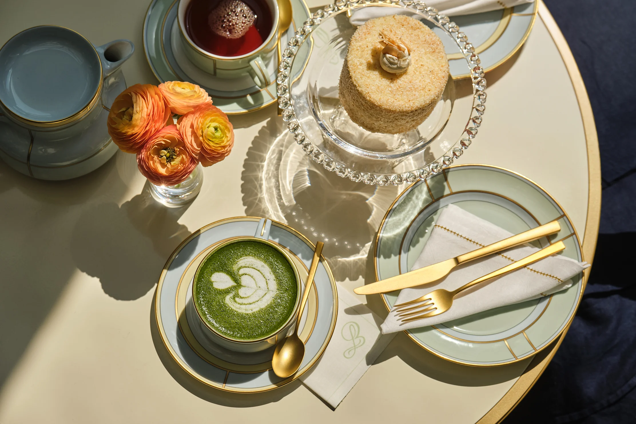 Elegant setup with matcha latte, tea, cake, orange flowers, and gold-trimmed china on white table
