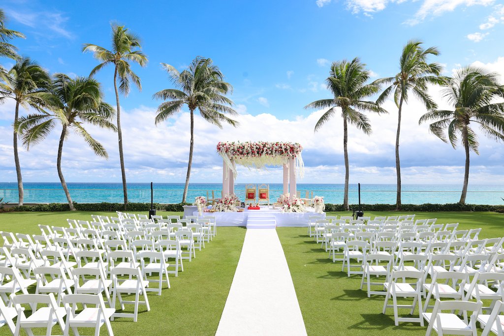 Beachfront wedding ceremony setup with white chairs, floral arch, palm trees, and ocean view.