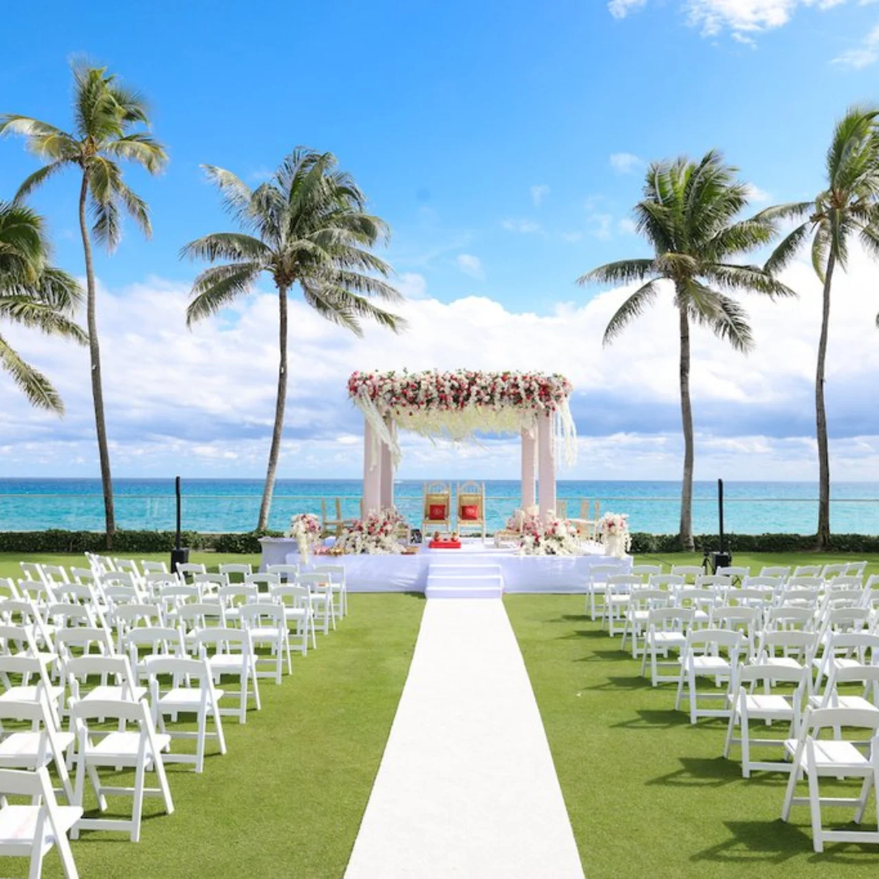 Beachfront wedding ceremony setup with white chairs, floral arch, palm trees, and ocean view.