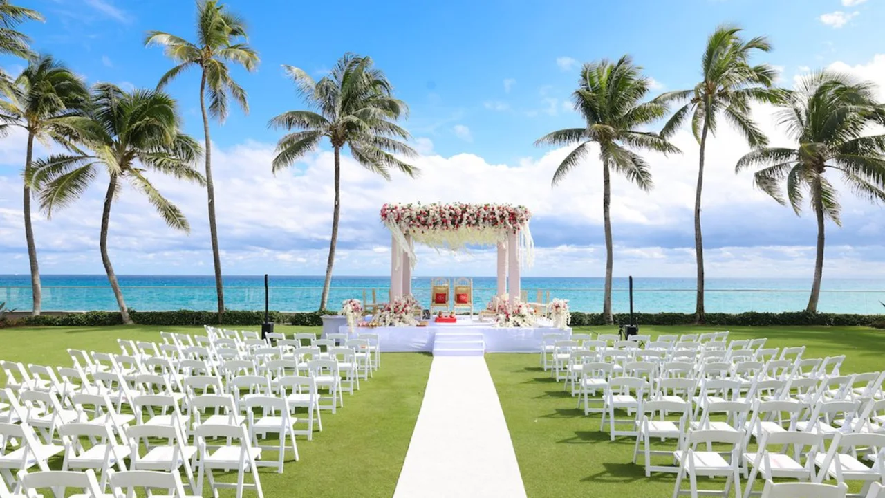 Beachfront wedding ceremony setup with white chairs, floral arch, palm trees, and ocean view.